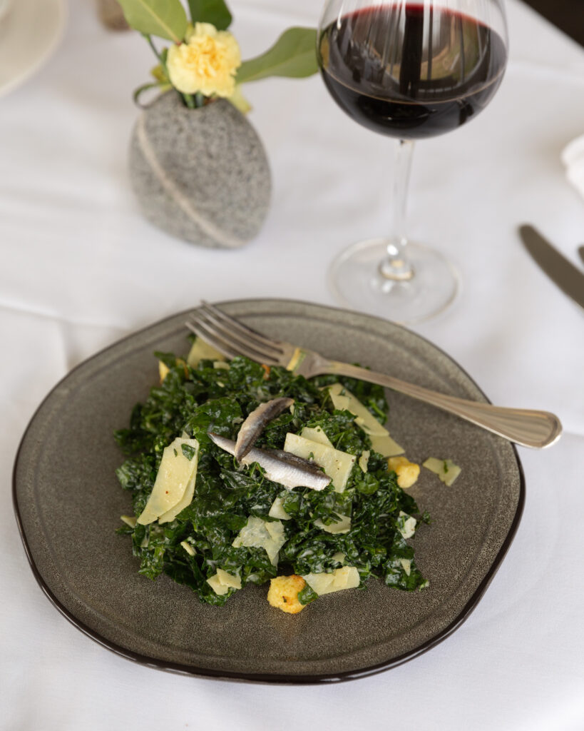 Detail photo of a glass of red wine next to a kale salad with anchovy filets on a stoneware plate  prepared by Chef Jon McCain, photographed by Laura Billingham Photography for River Towns Magazine.