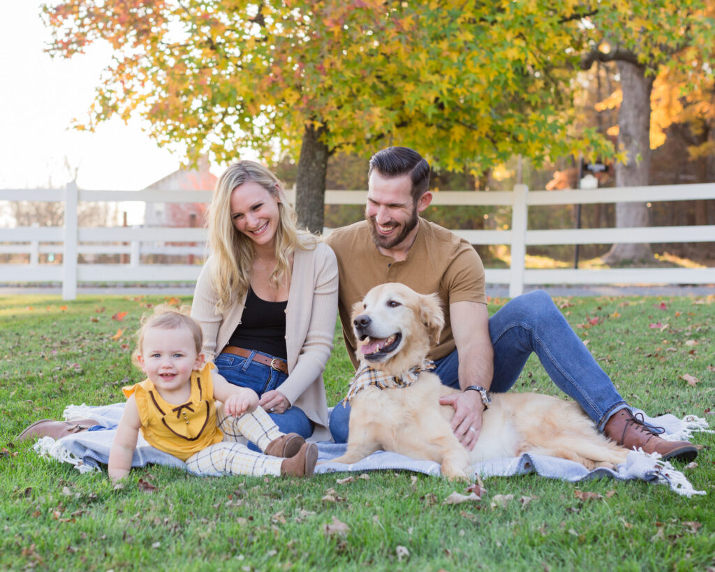 Family portrait of mom, dad, golden retriever dog and baby on a picnic blanket by white picket fence in the fall in their back yard in Whitehouse Station, NJ by Laura Billingham Photography