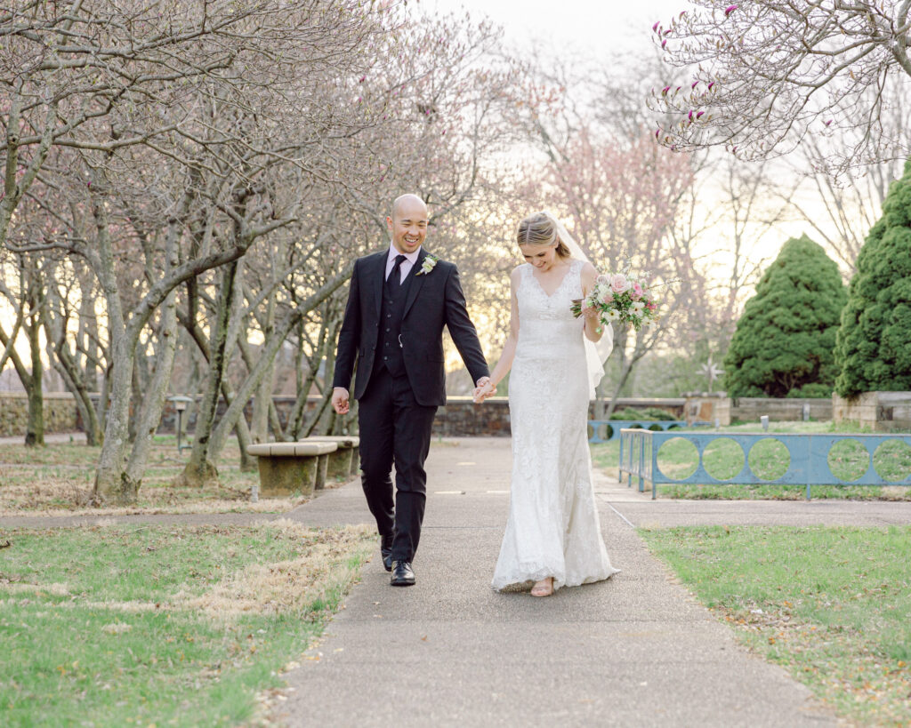 Elegant bride in a long lace gown and groom in a dark suit stroll hand in hand under blooming magnolia trees at sunset near the Bethlehem Area Public Library