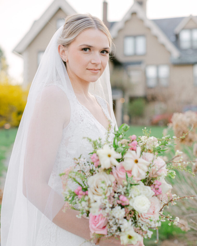 Bride wearing a veil holds a large luxurious spring bouquet of pink and ivory florals in front of the carriage house at the Sayre Mansion