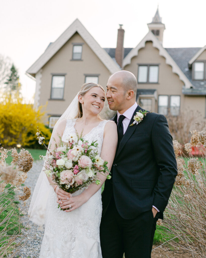 A bride in a sleeveless v-neck style lace gown holds a luxurious spring bouquet while a groom in a dark suit pulls her in to kiss her forehead in front of the carriage house at the Sayre Mansion