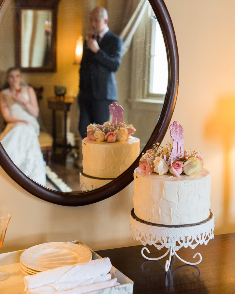 Elegant bride and groom, reflected in a mirror, sip champagne with a single tier wedding cake in the photo foreground