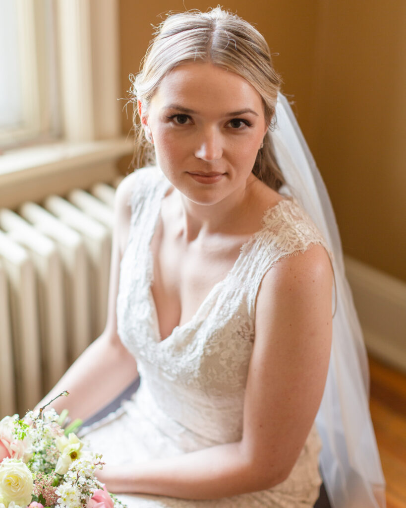 A bride wearing a v neck style lace gown is and a veil is seated with a lush bouquet of spring flowers in pins and ivories on her lap 