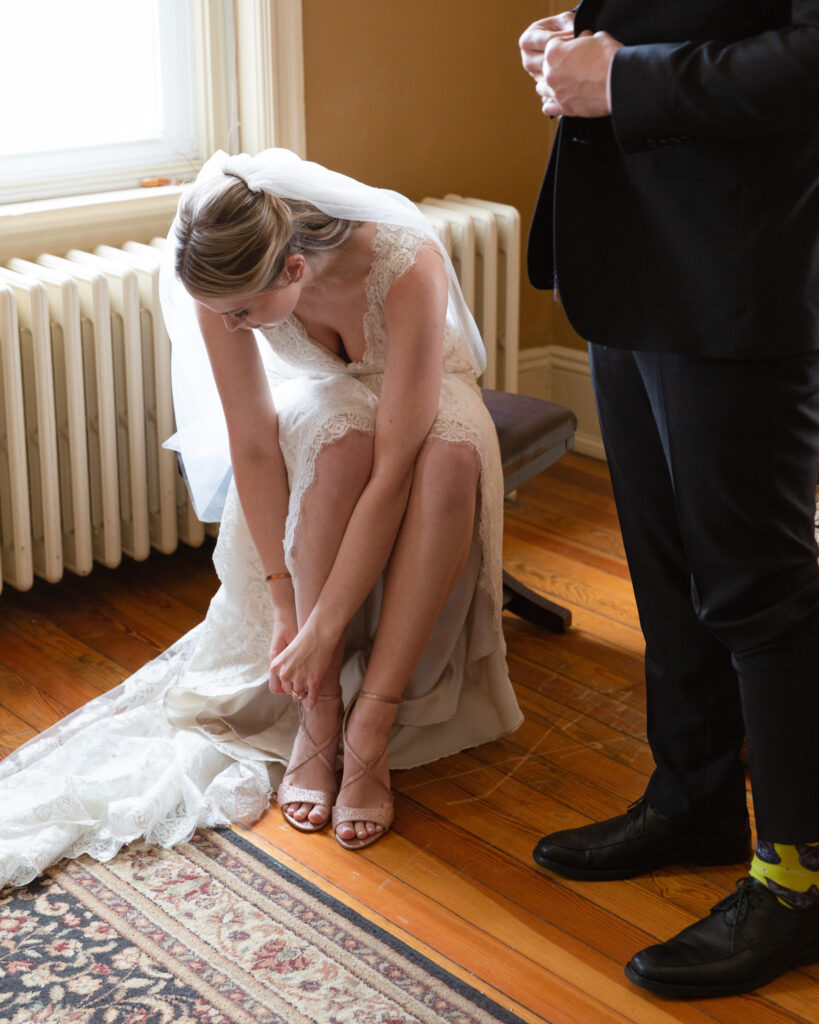 Bride seated on a bench buckles her heeled sandal while the groom in a dark suit stands next to her