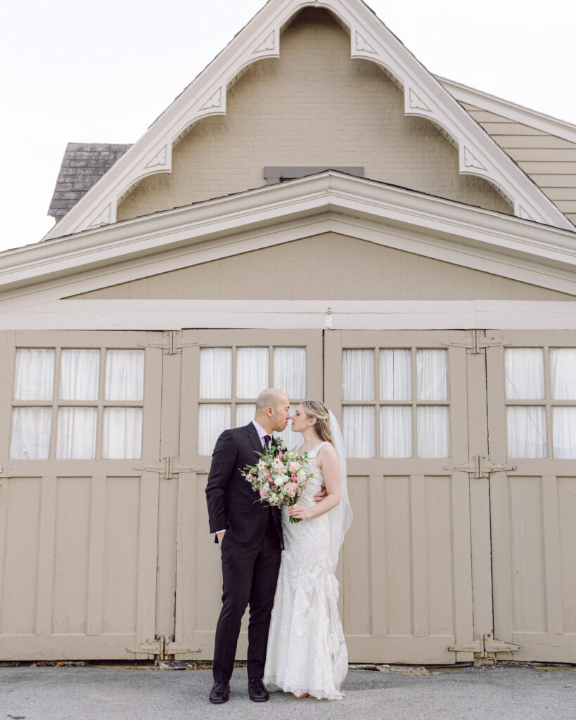 Bride with a veil and lace gown and groom in a dark suit kiss in front of an ornate vintage carriage barn