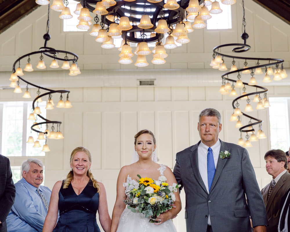 An elegant bride with a bouquet of sunflowers walks down the aisle with her mom and dad in the Coach House at the Ryland Inn in Whitehouse Station, NJ by Laura Billingham Photography