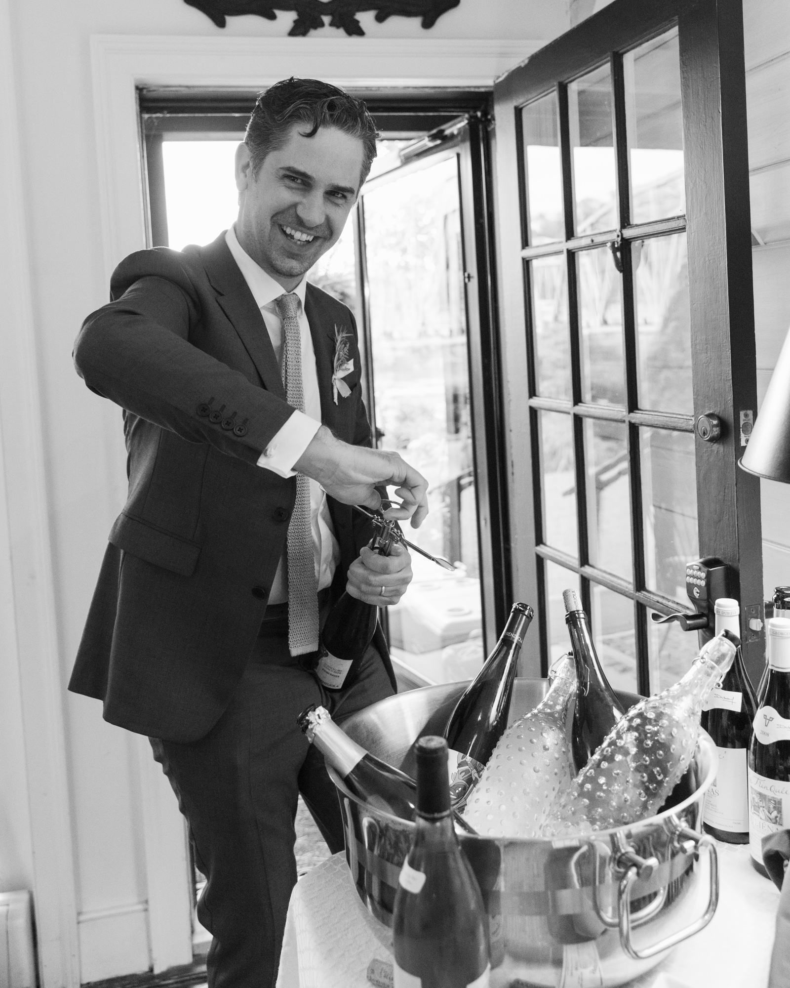 black and white photo of a stylish groom opening a bottle of champagne after his Bridgeton House on the Delaware wedding ceremony in Bucks County, PA