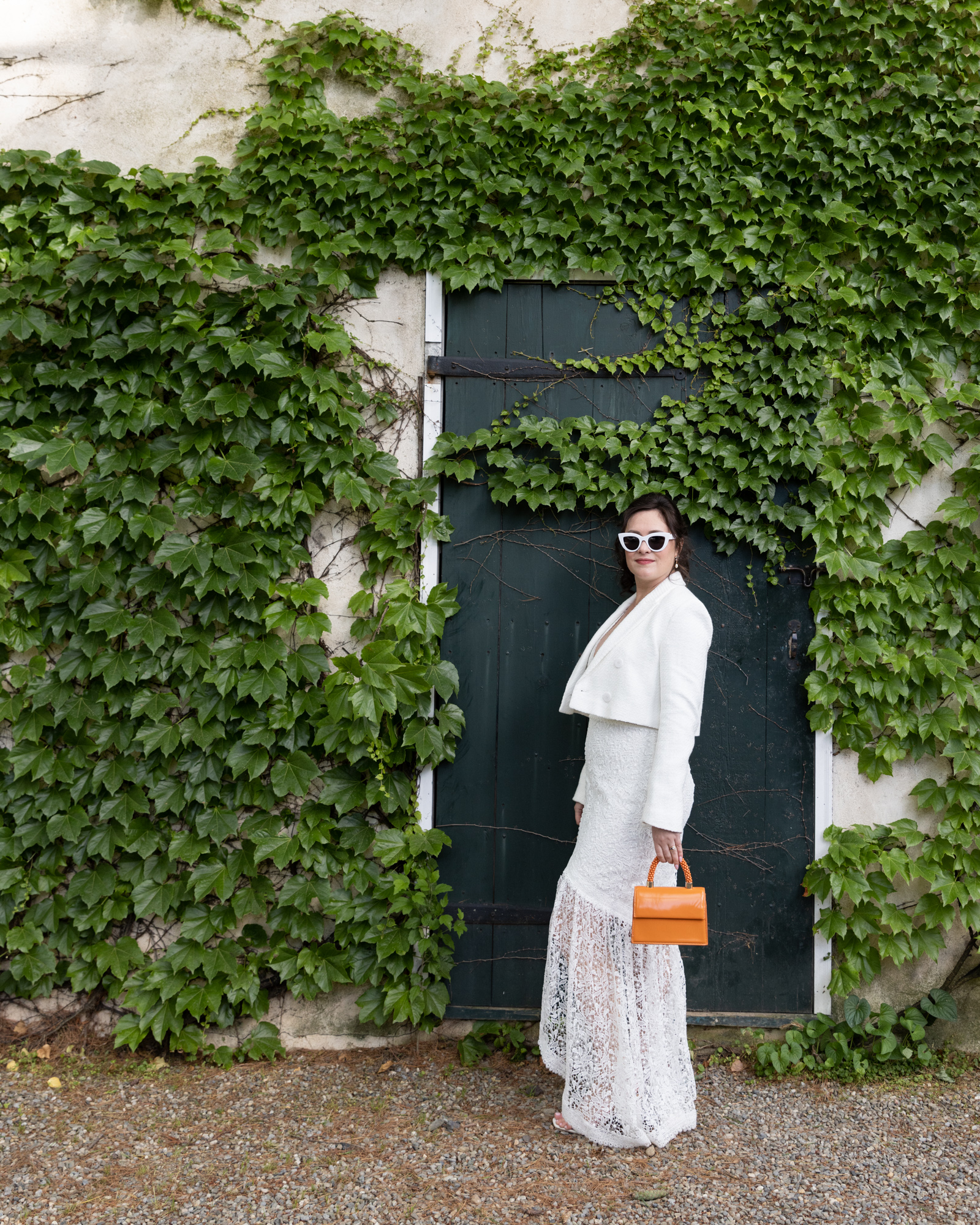 Cool bride poses wearing white cat eye sunglasses and orange purse in front of an ivy covered wall at Bridgeton House on the Delaware in Bucks County, PA