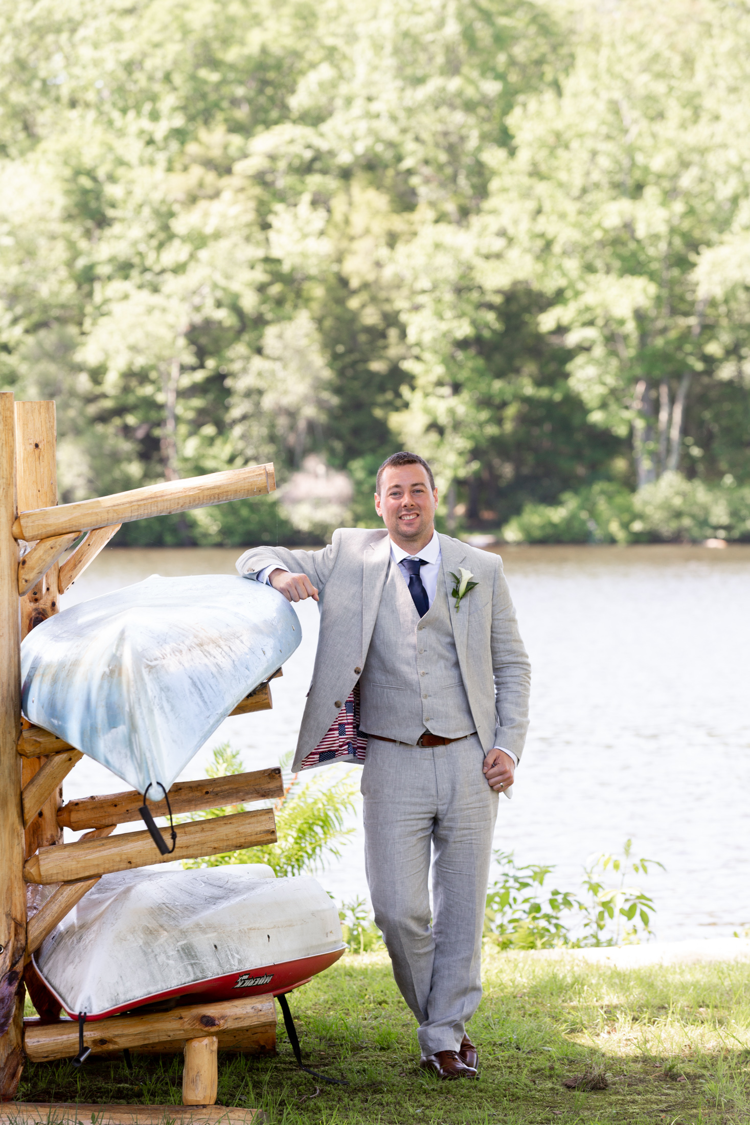 Elegant groom in a linen suit leans on a canoes stacked near the lake at Mountain Springs Lake Resort in the Poconos in Reeders, PA