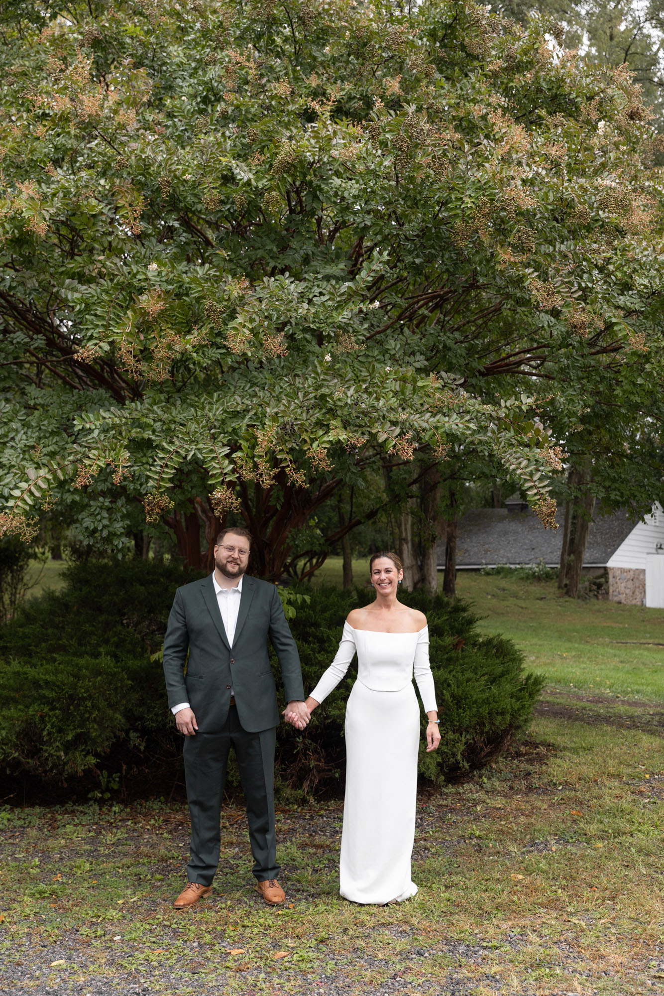 An elegant bride in an elegant modern sheath dress and groom in a dark green suit hold hands after their wedding ceremony at Chimney Hill Estate Inn in Lambertville, NJ