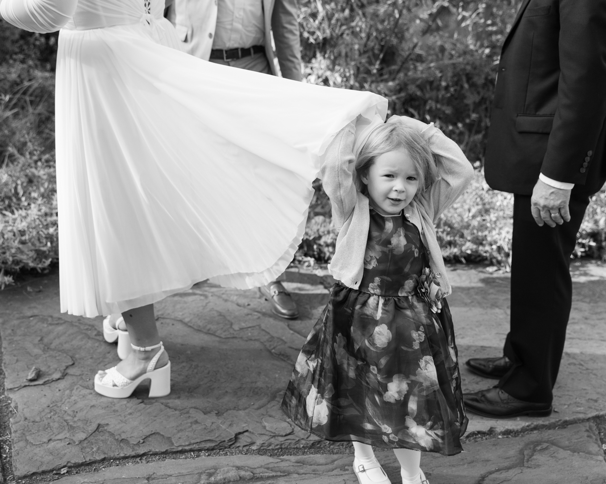 black and white candid photo of a little girl playing hide and seek with the back of a bride's chiffon skirt