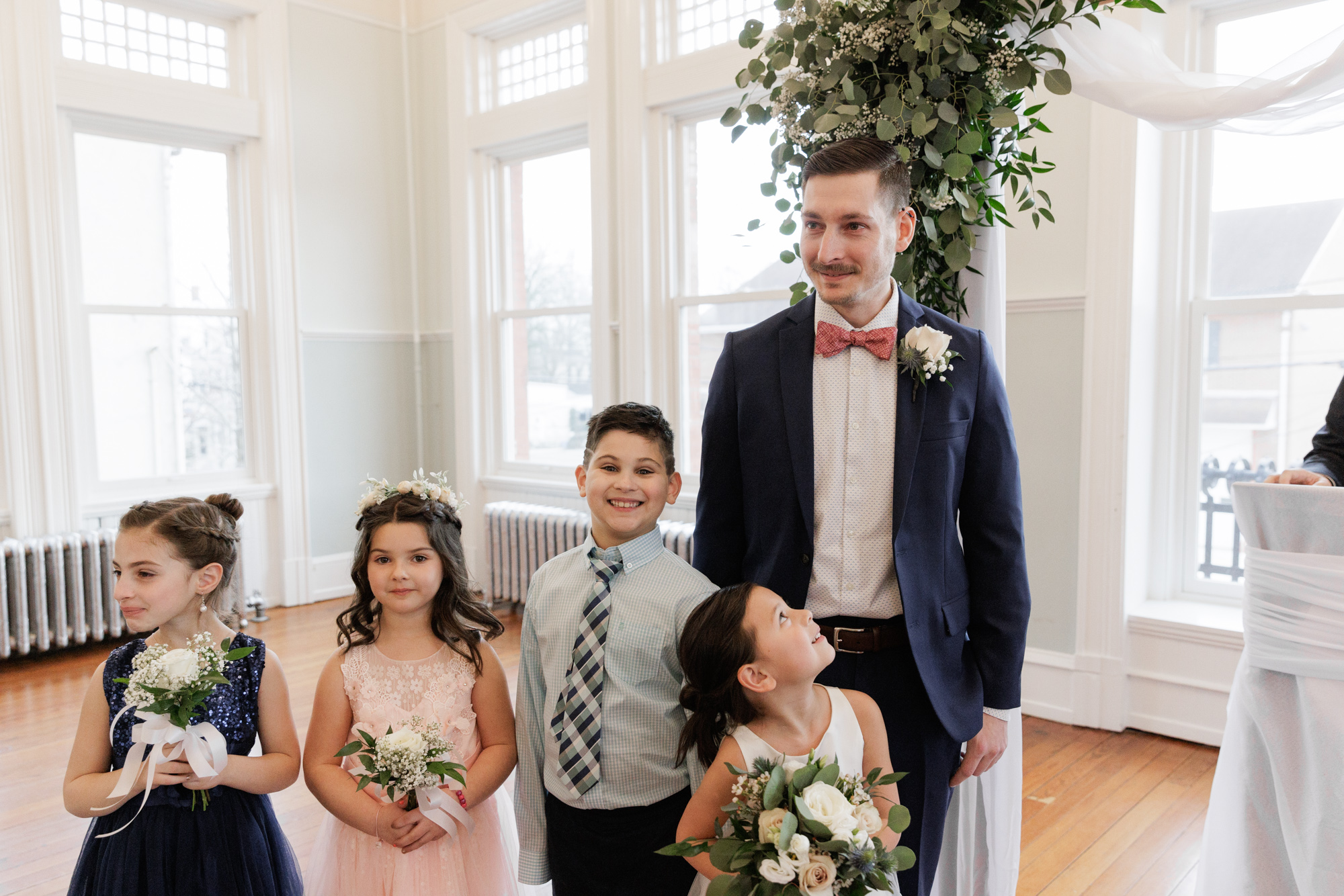 Elegant groom waits for the bride while surrounded by children in the wedding party at the altar during a wedding ceremony at Old City Hall in Bordentown, NJ 