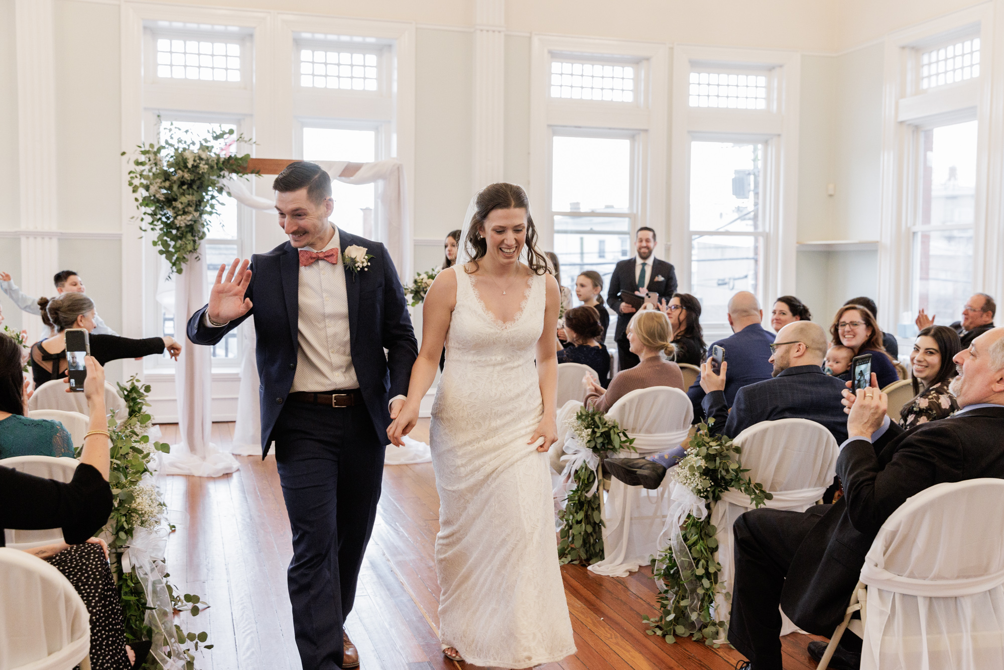 Elegant groom waves to guests as he and bride walk down the aisle after their ceremony at historic Old City Hall in Bordentown, NJ