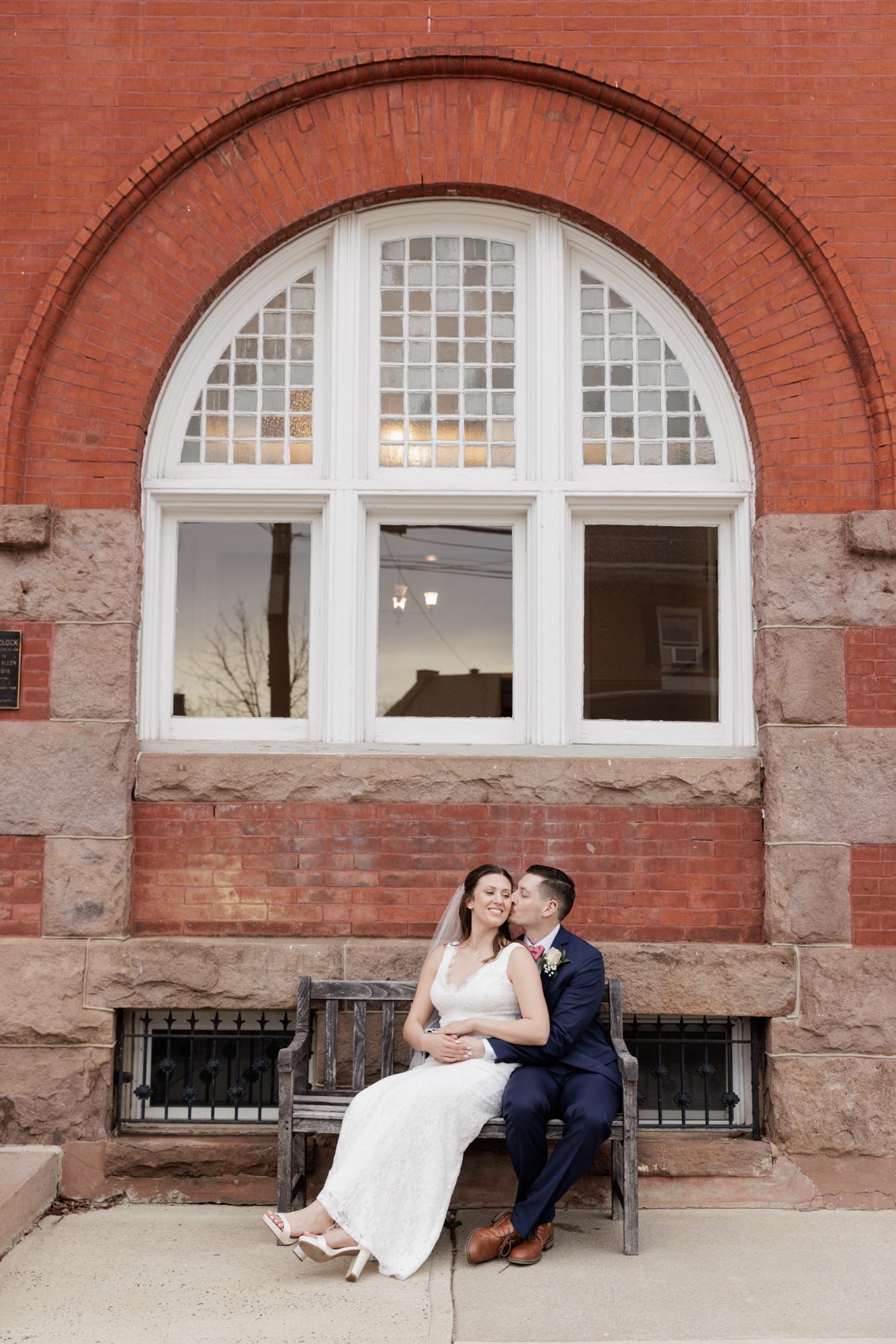 Elegant bride and groom embrace on a bench under an historic arched window at Old City Hall in Bordentown, NJ