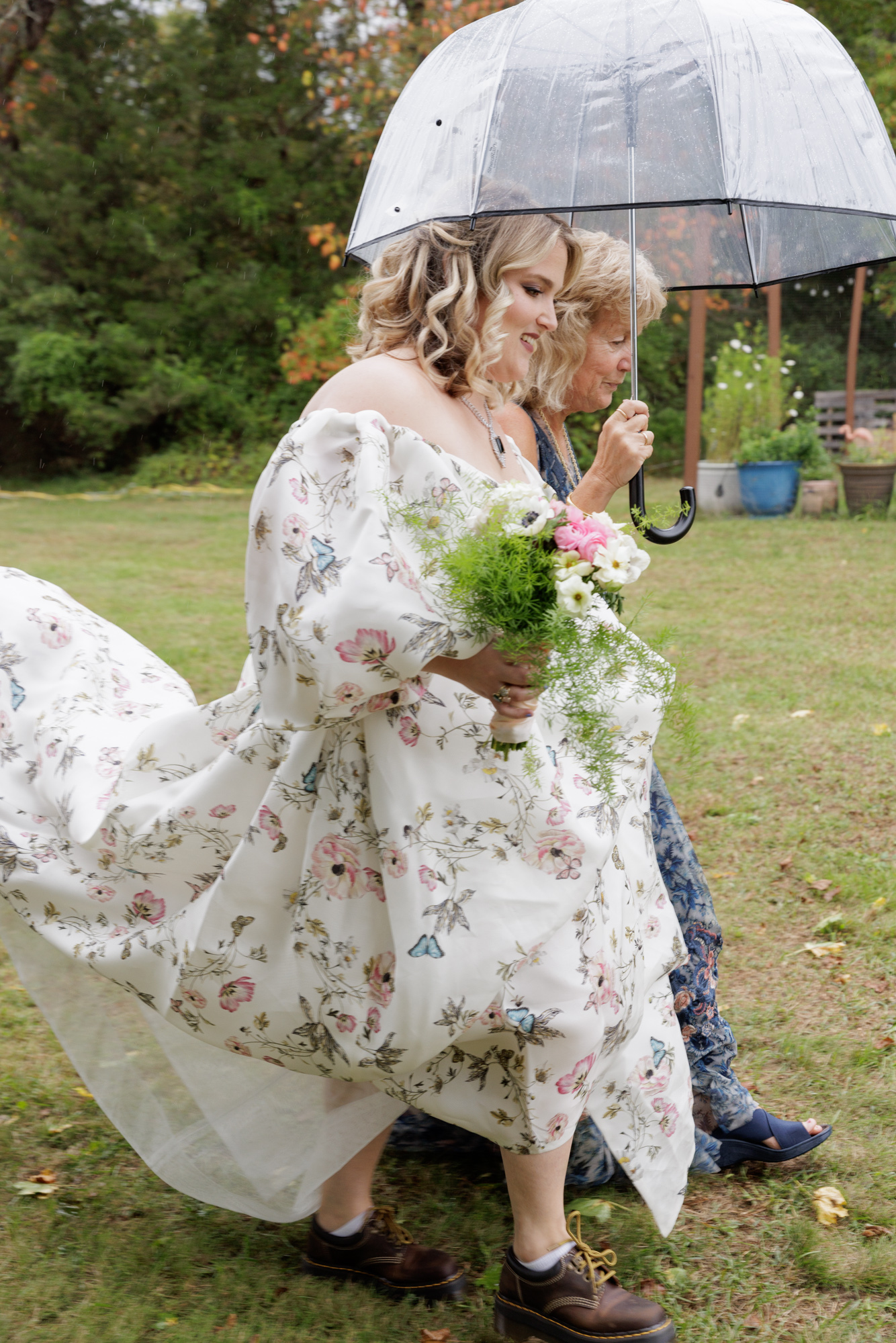 Mother of the bride holds a clear umbrella over the head of a bride in a long flowing floral Monique Lhullier gown as they process toward a backyard wedding ceremony in NJ