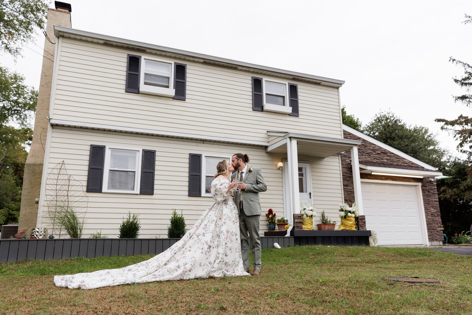 Bride in a full, long floral Monique Lluillier gown and groom in a light colored suit in a formal pose in front of their two story house in New Jersey