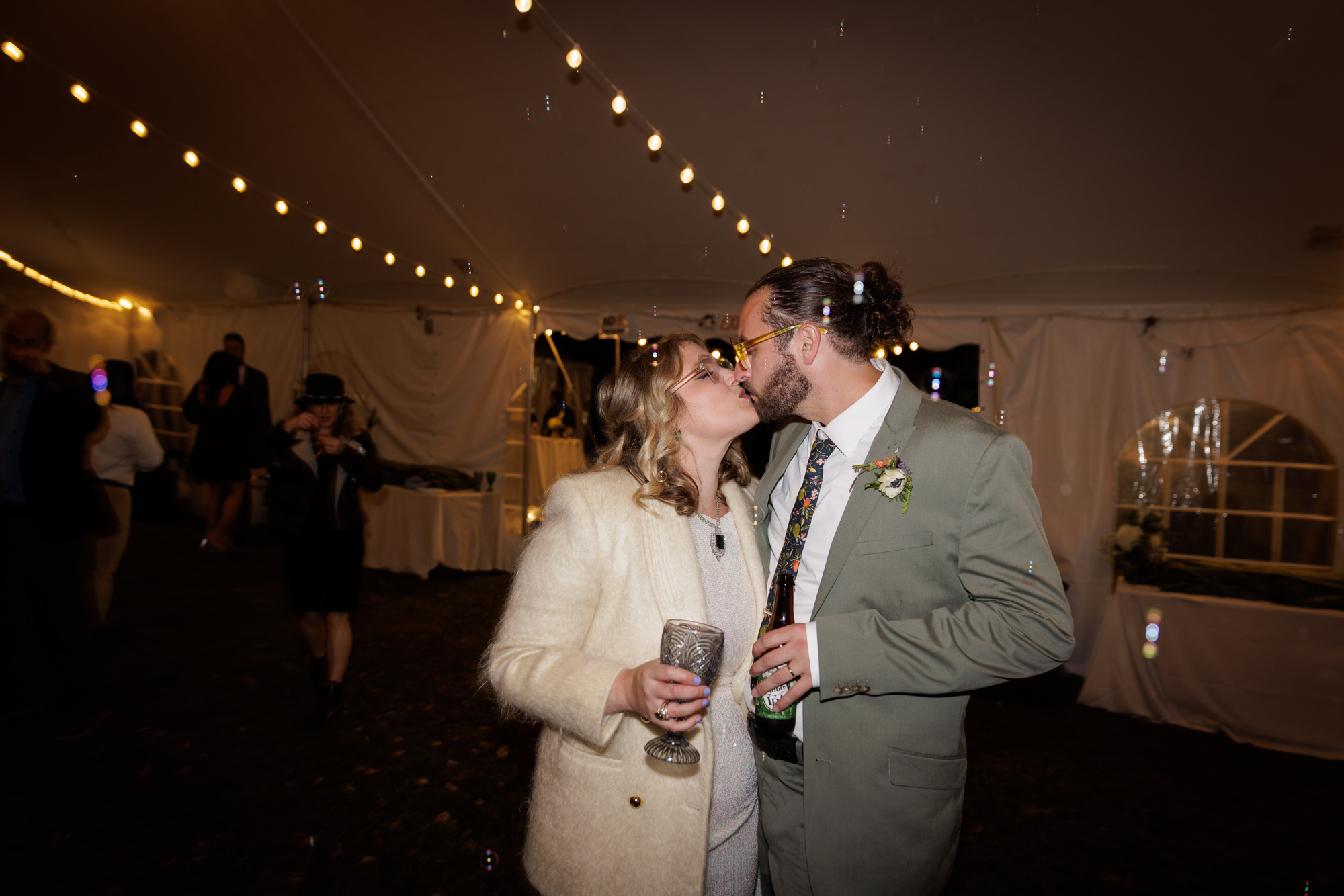a bride in a white mohair double breasted blazer and sequined wedding dress kisses a groom in a sage green jacket and floral tie in their tented recption an at home wedding reception in New Jersey. The flash wedding photo has some motion blur of bubbles guests have blown at them and strings of cafe lights at the ceiling.