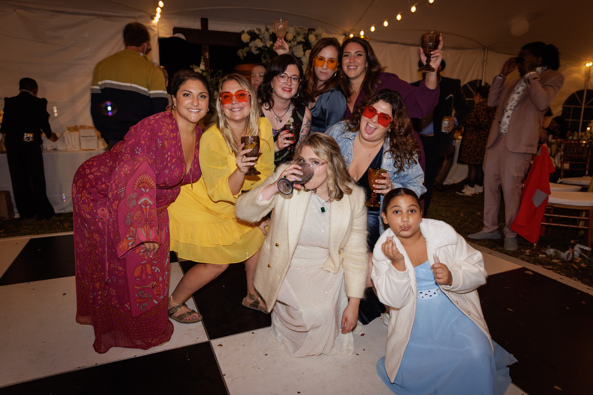 a crowd of young women in colorful dresses and brightly colored sunglasses gather around a bride in a white mohair blaser and sequined dress on a black and white checkered dance floor at a tented at home wedding in NJ