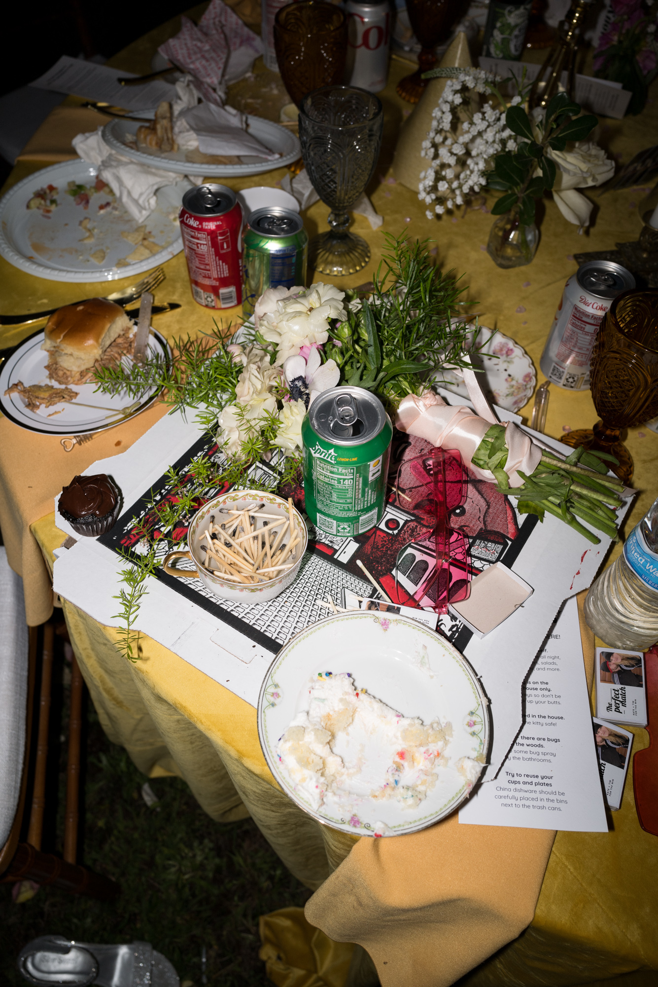 a messy, colorful tablecloth piled high with a pizza box, soda can and wedding bouquet at the end of an at home wedding reception in New Jersey