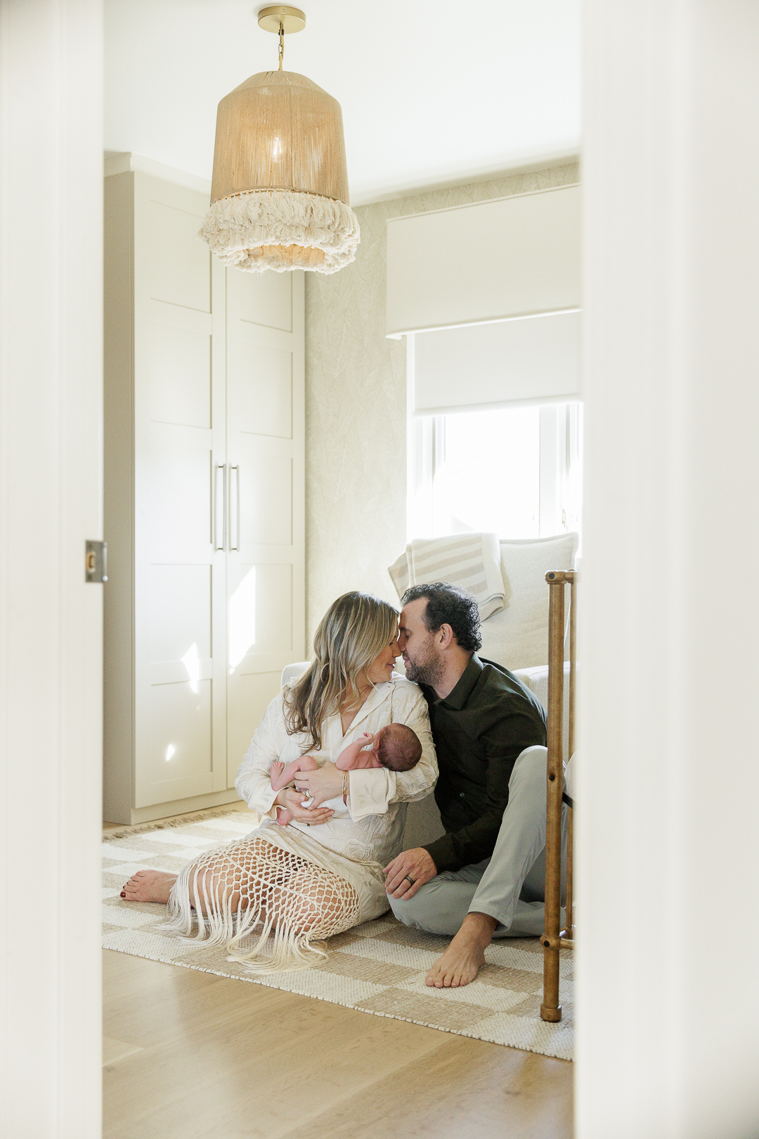 A mother and father cuddle a newborn baby while sitting casually on the floor of a modern neutral nursery