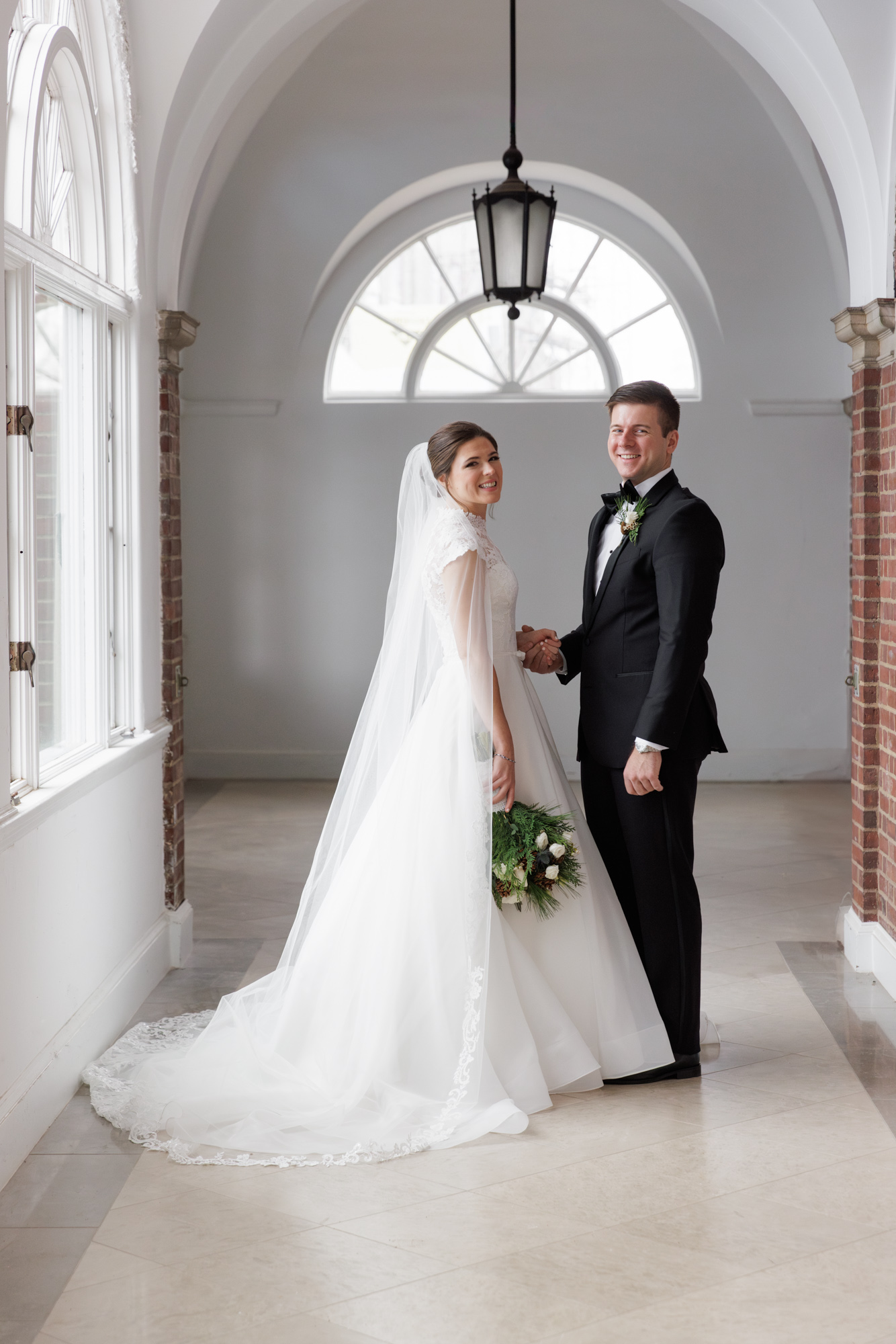 An elegant bride in a long white gown and holding a bouquet at her side holds the hand of a groom in black tie attire in an arched white hallway at the Mansion at Natirar in Peapack and Gladstone NJ