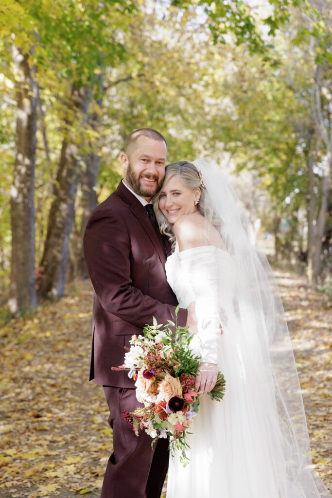 An elegant groom in a burgundy suit and a bride in a long white chiffon gown with a veil snuggle and smile at the camera. They are standing on a path next to the Delaware River in Frenchtown, NJ and the trees around them are turning gold in late fall.