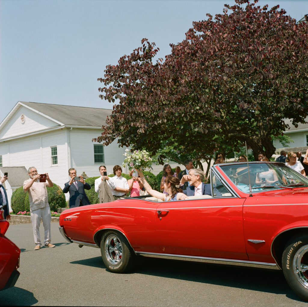 A bride and groom turn to wave at guests while riding in a vintage red convertible getaway car