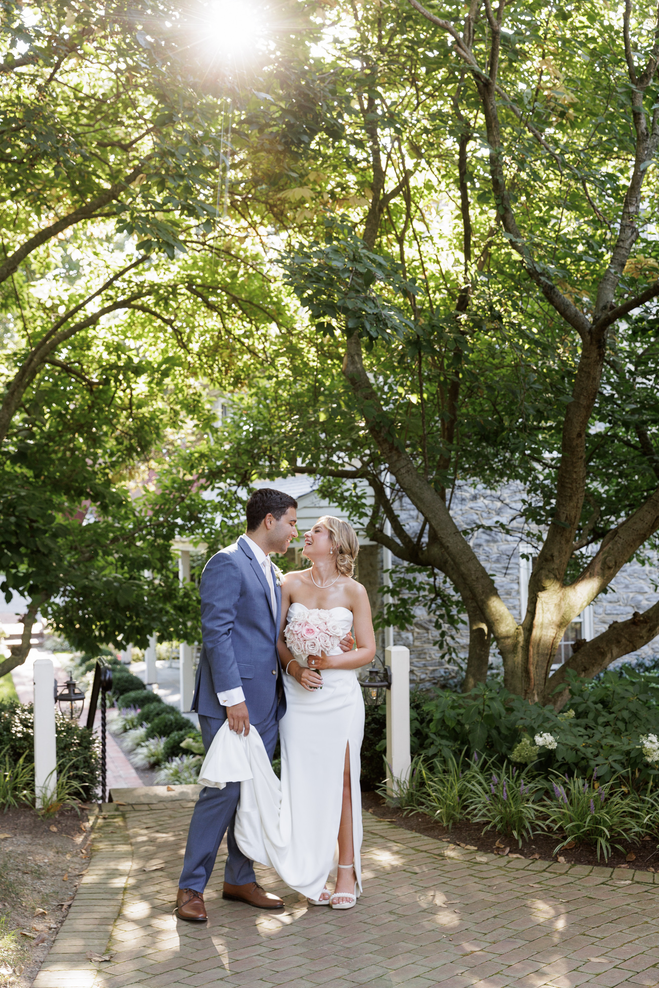 A bride in an elegant sleeveless gown and a groom in a blue suit are backlit by bright sunshine while they steal a moment together