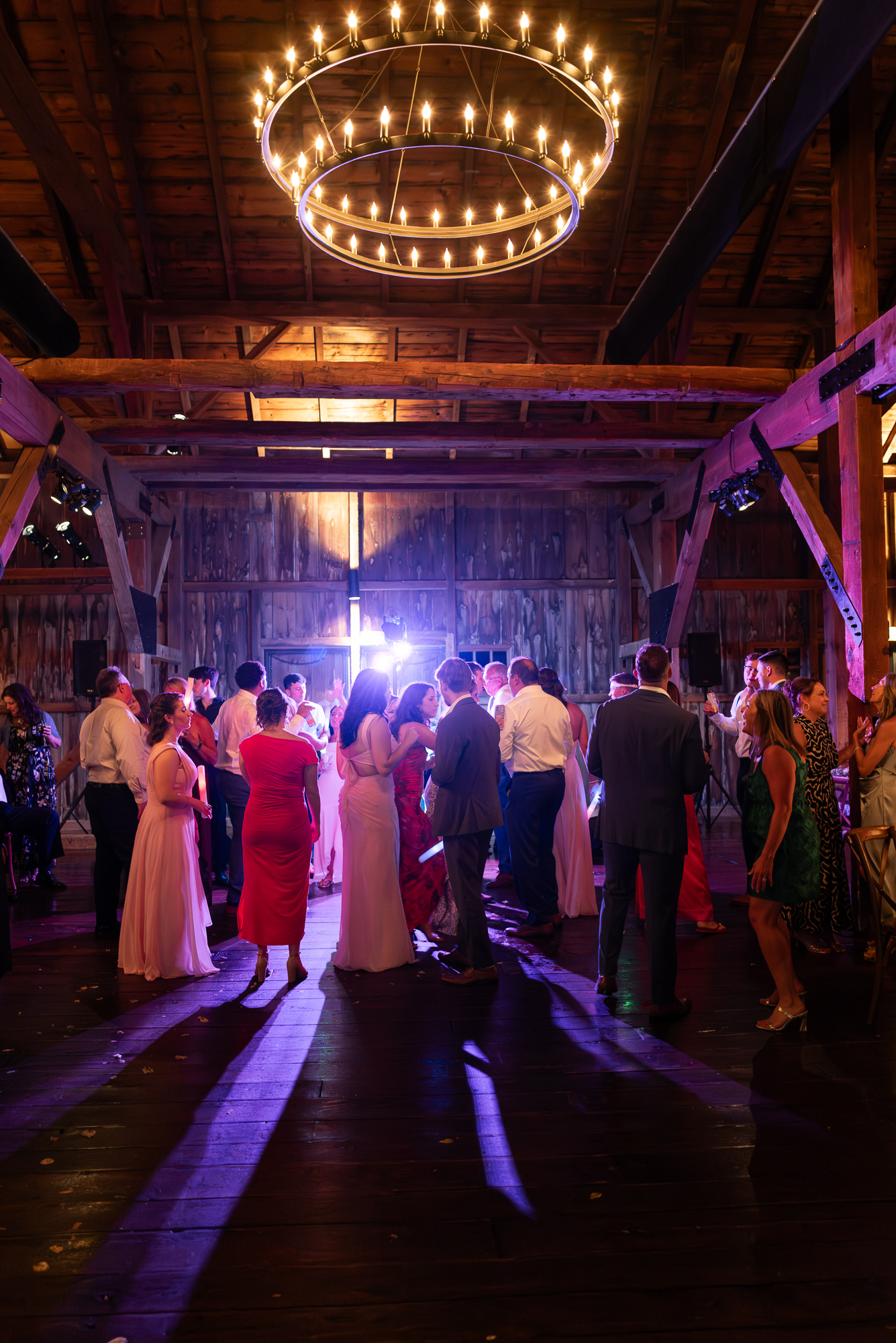 Wedding dance floor under a large two tiered rustic chandelier is full of guests backlit by disco lights