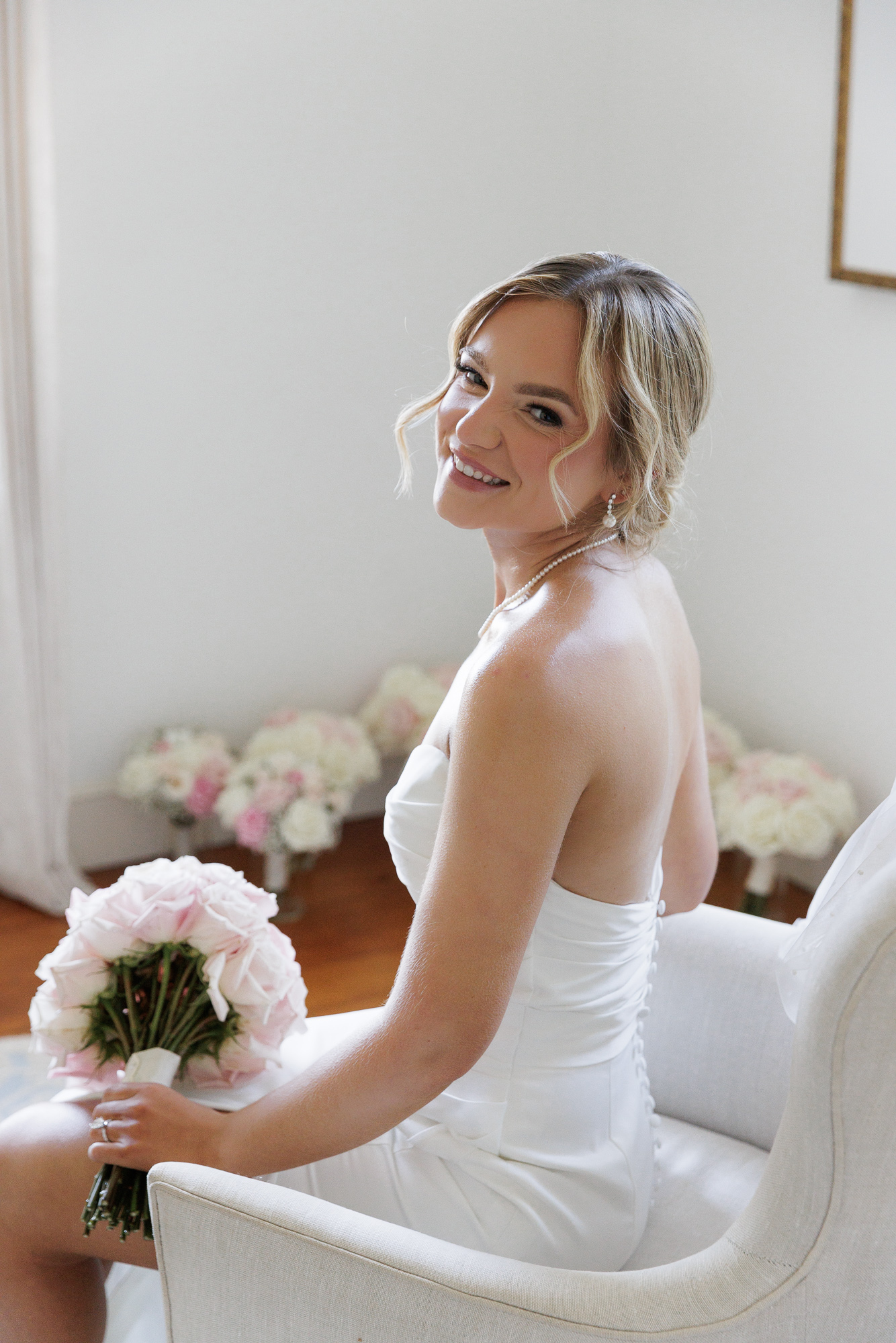 A bride in an elegant sleeveless gown sits in a linen upholstered arm chair, looking over her shoulder while a bouquet of pink roses rests on her lap