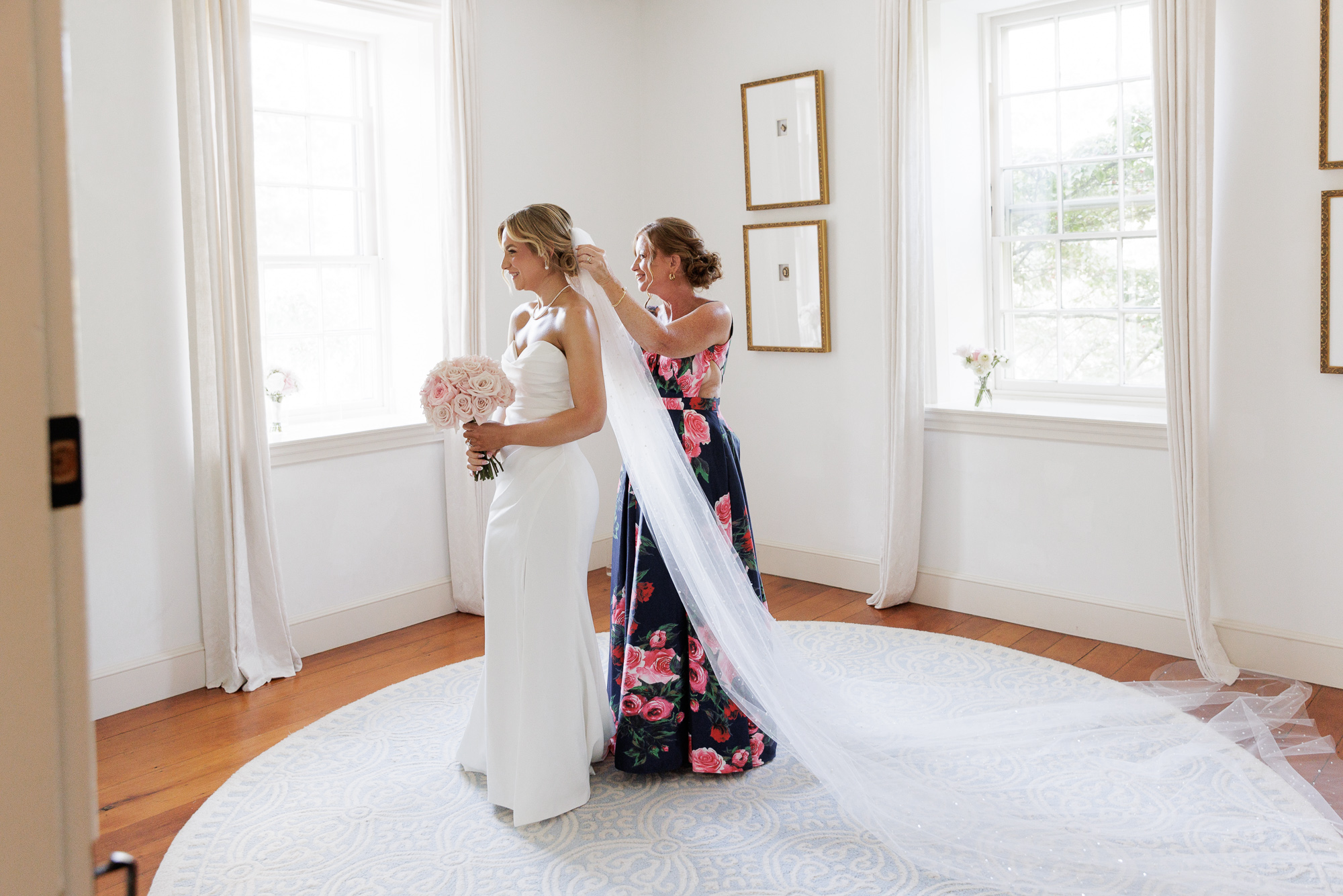 Bride in an elegant white gown stands in front of a floor to ceiling mirror while her mom places a cathedral length veil on her before a wedding ceremony at Farm at Eagles Ridge in Lancaster, PA