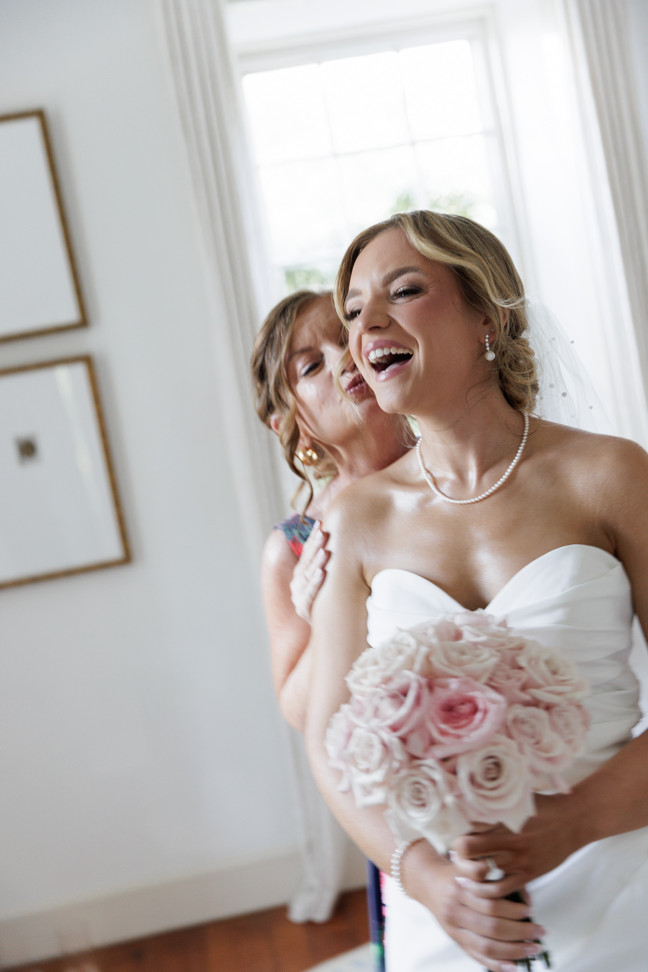 A bride in an elegant sleeveless gown holds a lush bouquet of soft pink roses and laughs while her mom standing behind her kisses her cheek