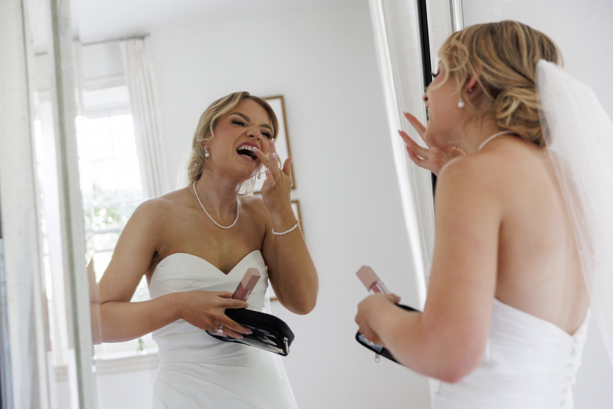A bride in an elegant sleeveless gown looks into a large mirror to wipe lipstick off her teeth