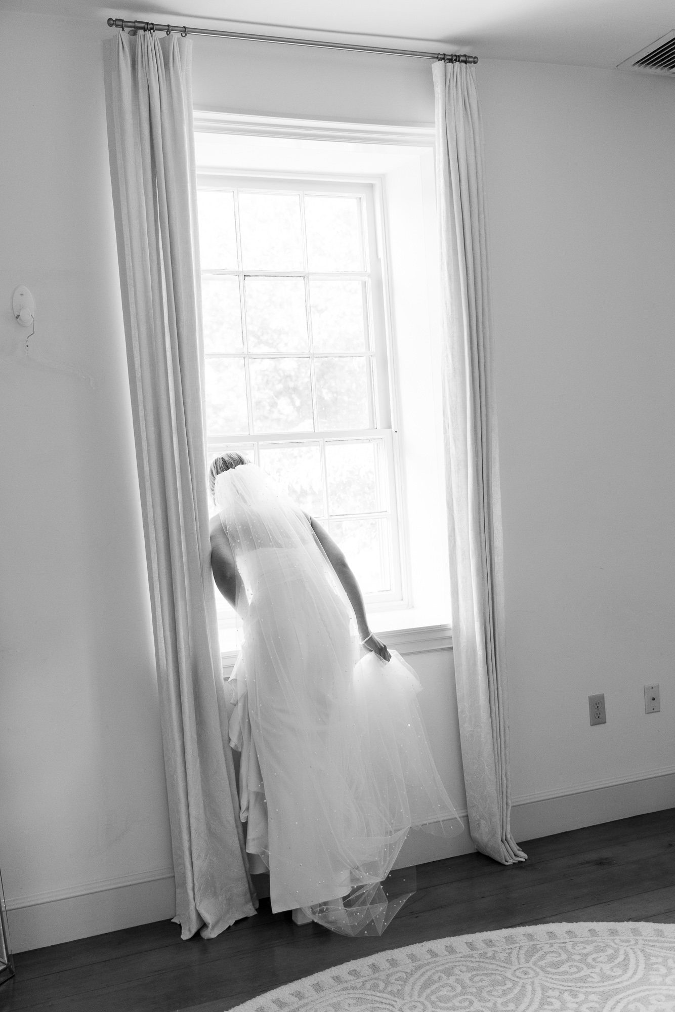 A bride in a long white sleeveless gown and veil looks out the window expectantly at arriving wedding guests before her wedding at Farm at Eagles Ridge in Lancaster, PA