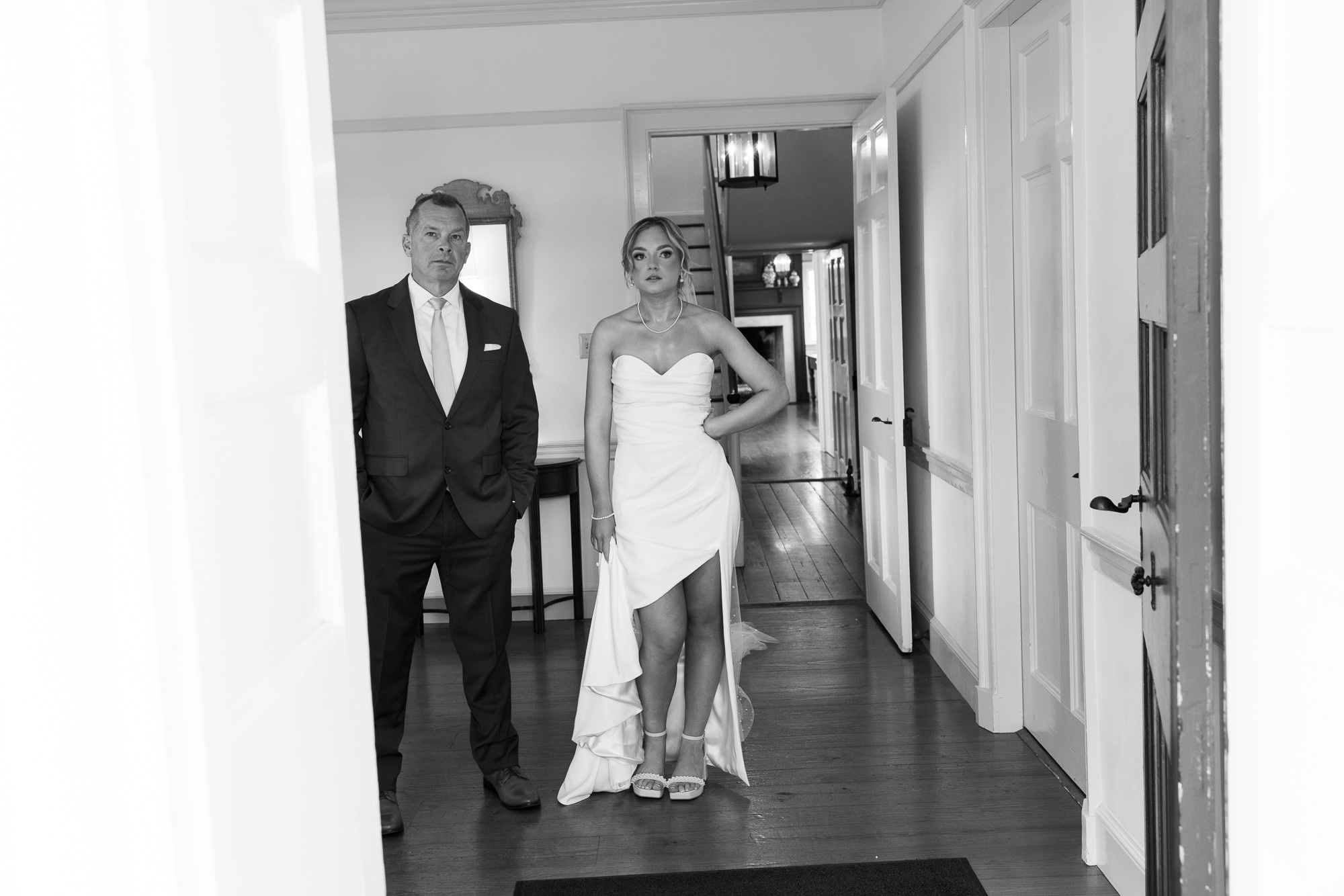 A bride in an elegant sleeveless wedding gown holds the hem of her dress while casually standing next to her dad in a doorway at Farm at Eagles Ridge in Lancaster, PA