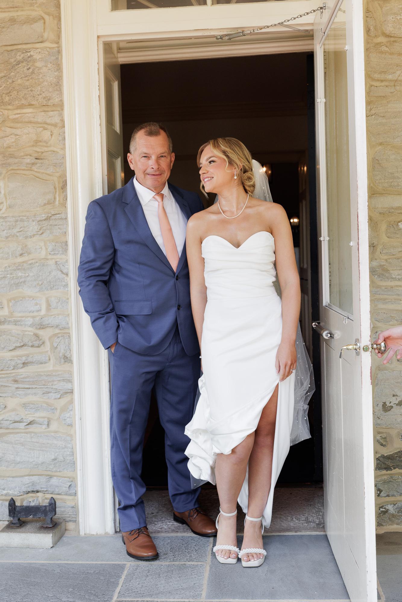 A bride in an elegant sleeveless wedding gown holds the hem of her dress while casually standing next to her dad in a doorway at Farm at Eagles Ridge in Lancaster, PA