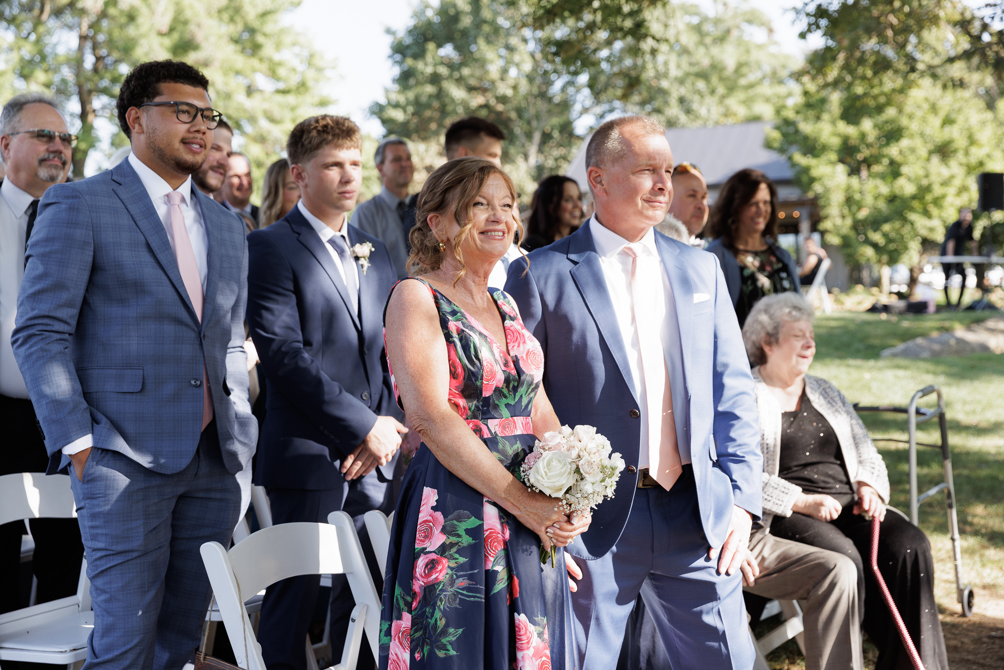 A smiling mother of the bride wearing a sleeveless navy floral gown and holds a small bouquet of flowers next to the smiling father of the bride during a wedding ceremony at Farm at Eagles Ridge in Lancaster, PA
