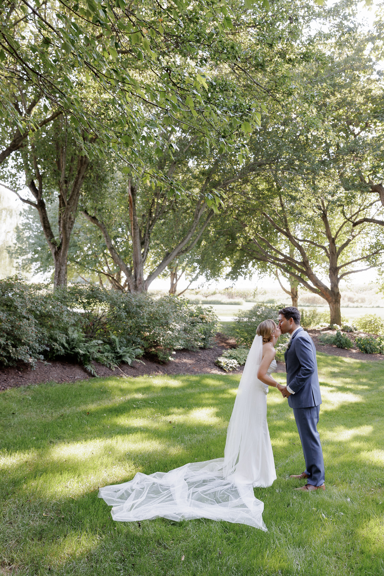 A bride in an elegant sleeveless gown and cathedral length veil holds hands with groom in a blue suit as they lean toward each other to kiss on a sun dappled lawn at Farm at Eagles Ridge in Lancaster, PA