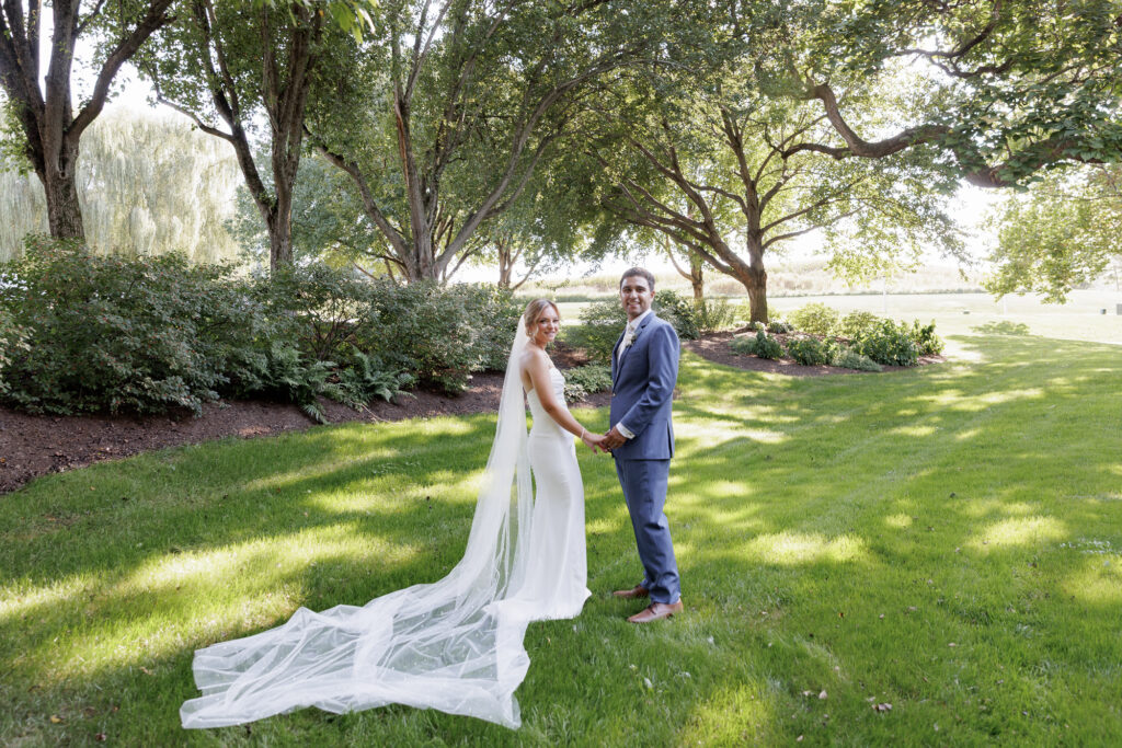 A bride in a cathedral length veil and white off the shoulder gown holds hands with a groom in a blue suit on a sunlight dappled lawn at Farm at Eagles Ridge in Lancaster, PA