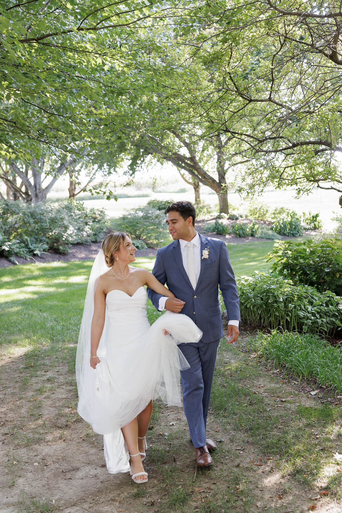 Bride in an elegant sleeveless gown and long veil walks closely with groom in a blue suit at Farm at Eagles Ridge in Lancaster, PA