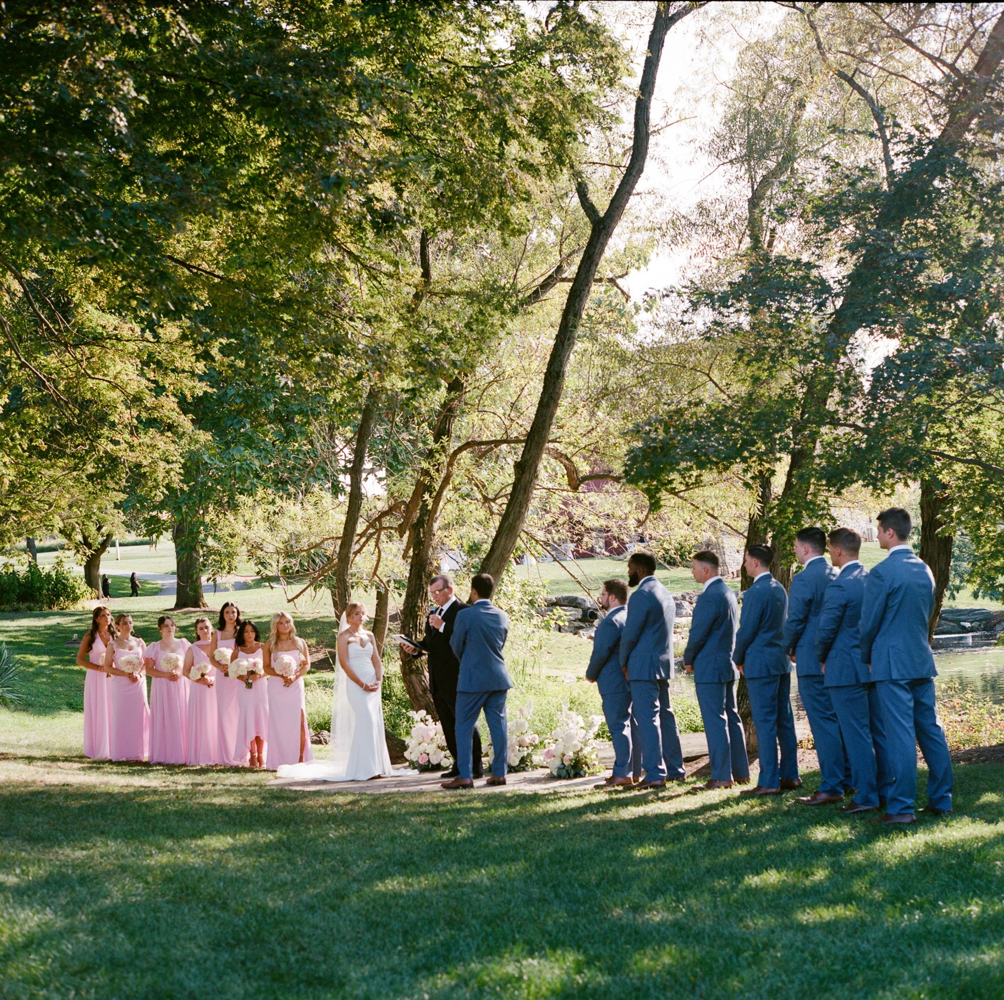 Wedding party of bridesmaids in soft pink chiffon gowns and groomsmen in blue suits stand with bride and groom during a wedding ceremony at the Dock at Farm at Eagles Ridge in Lancaster, PA