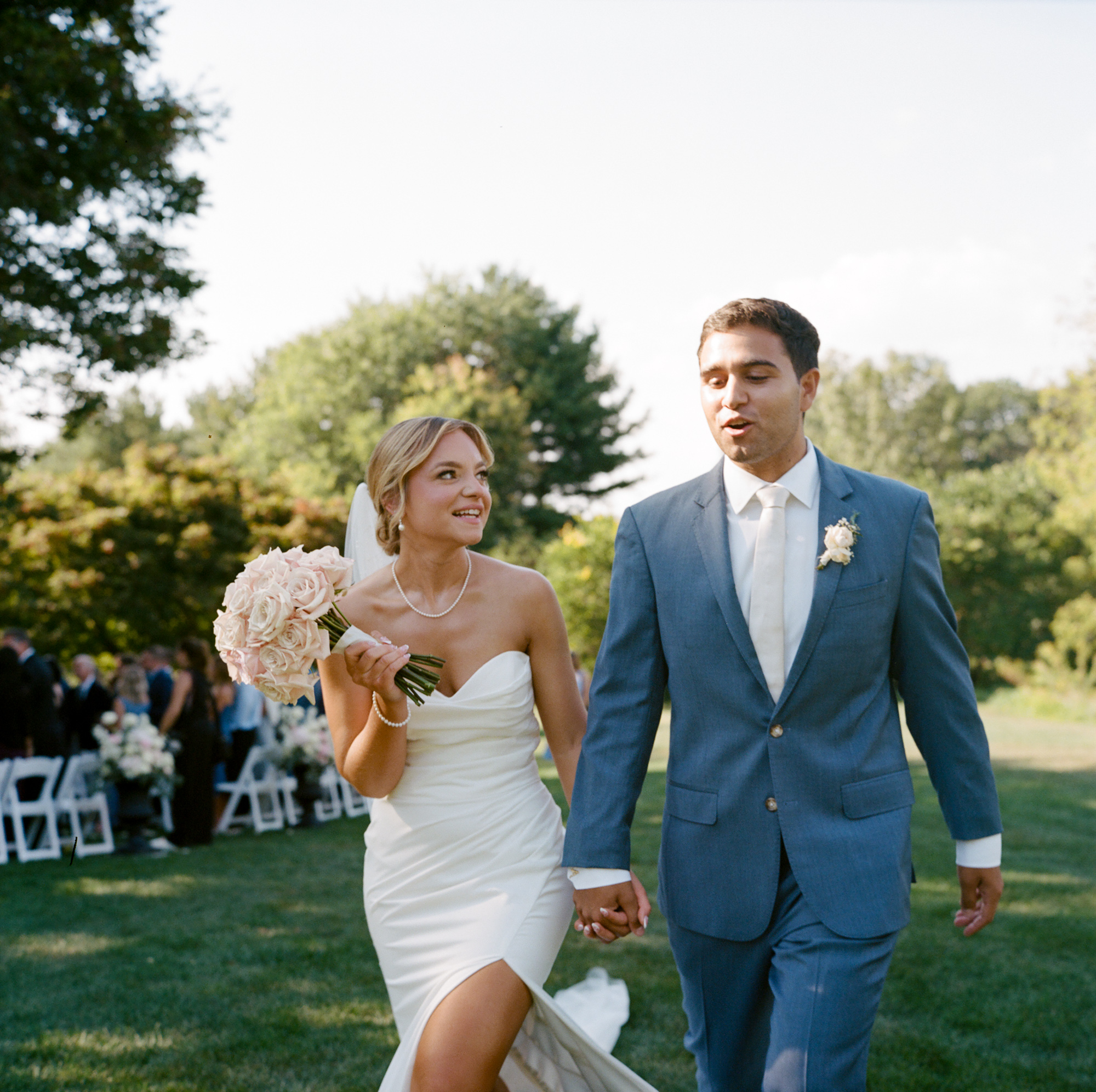 Bride with her bouquet of roses over her shoulder and groom walk hand in hand away from their wedding ceremony at Farm at Eagles Ridge in Lancaster, PA