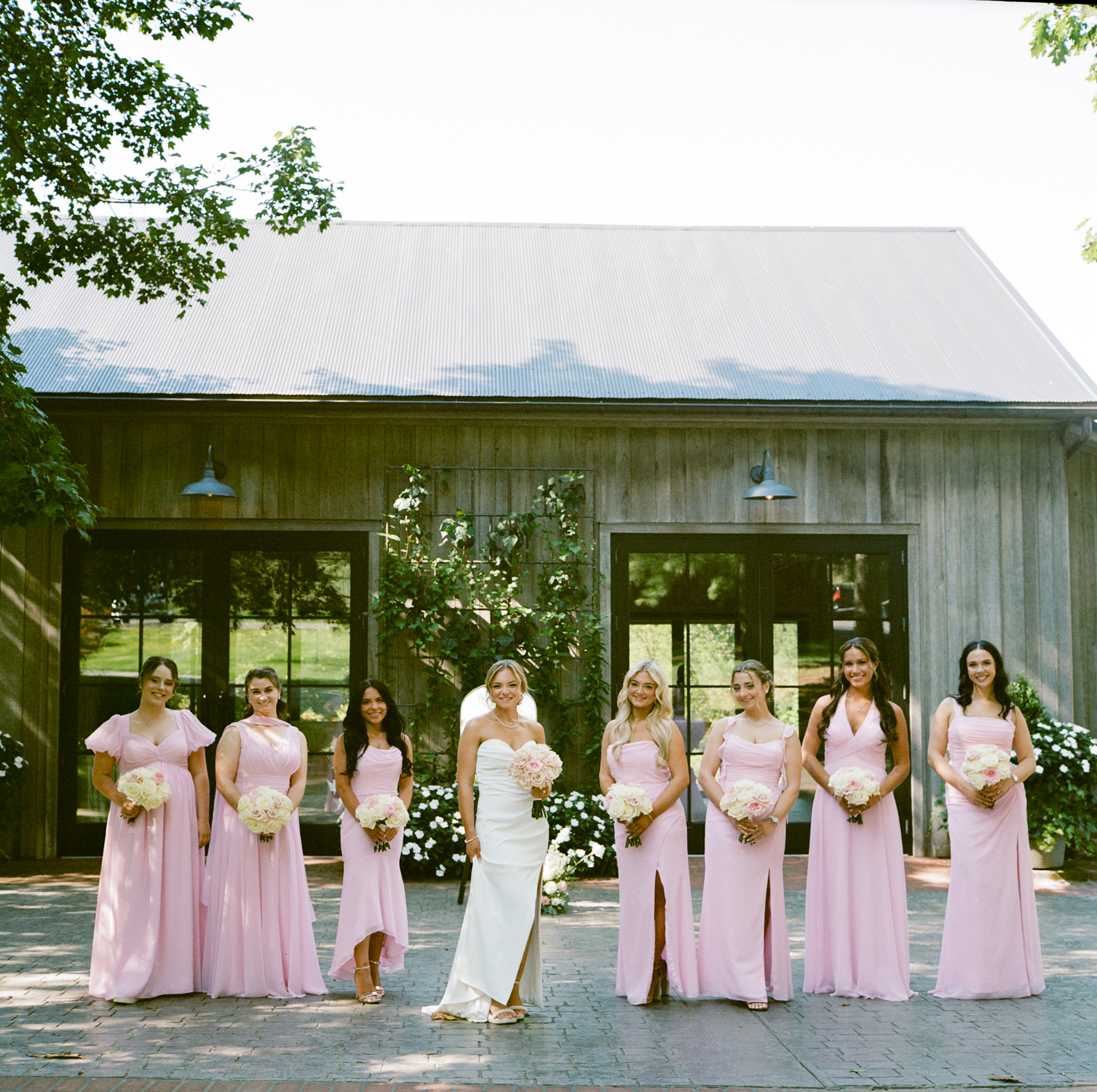 Bride in an elegant sleevelses gown strikes a cool pose while surrounded by her bridesmaids all wearing soft pink gowns in front of the Carriage House at Farm at Eagles Ridge in Lancaster, PA