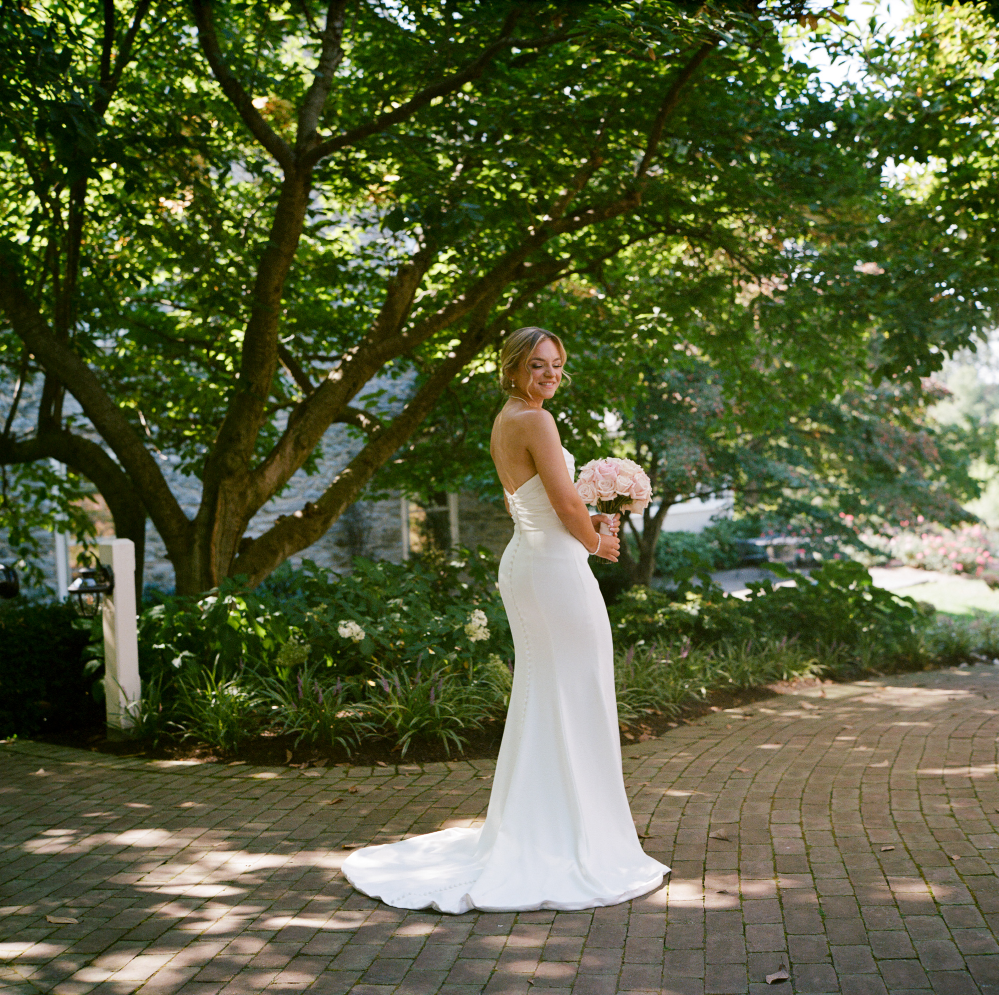 Bride in an elegant long sleeveless gown holding a bouquet of soft pink roses stands on a brick walkway under trees at Farm at Eagles Ridge in Lancaster, PA