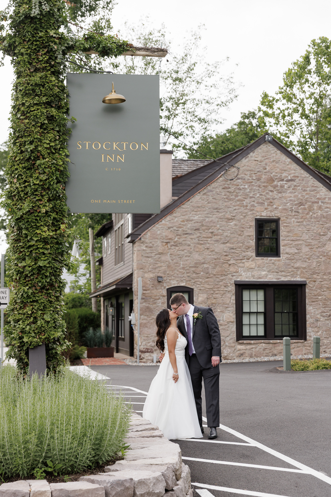 A bride in a long summery gown kisses a groom in a dark suit under the ivy covered sign of the Stockton Inn in Stockton NJ
