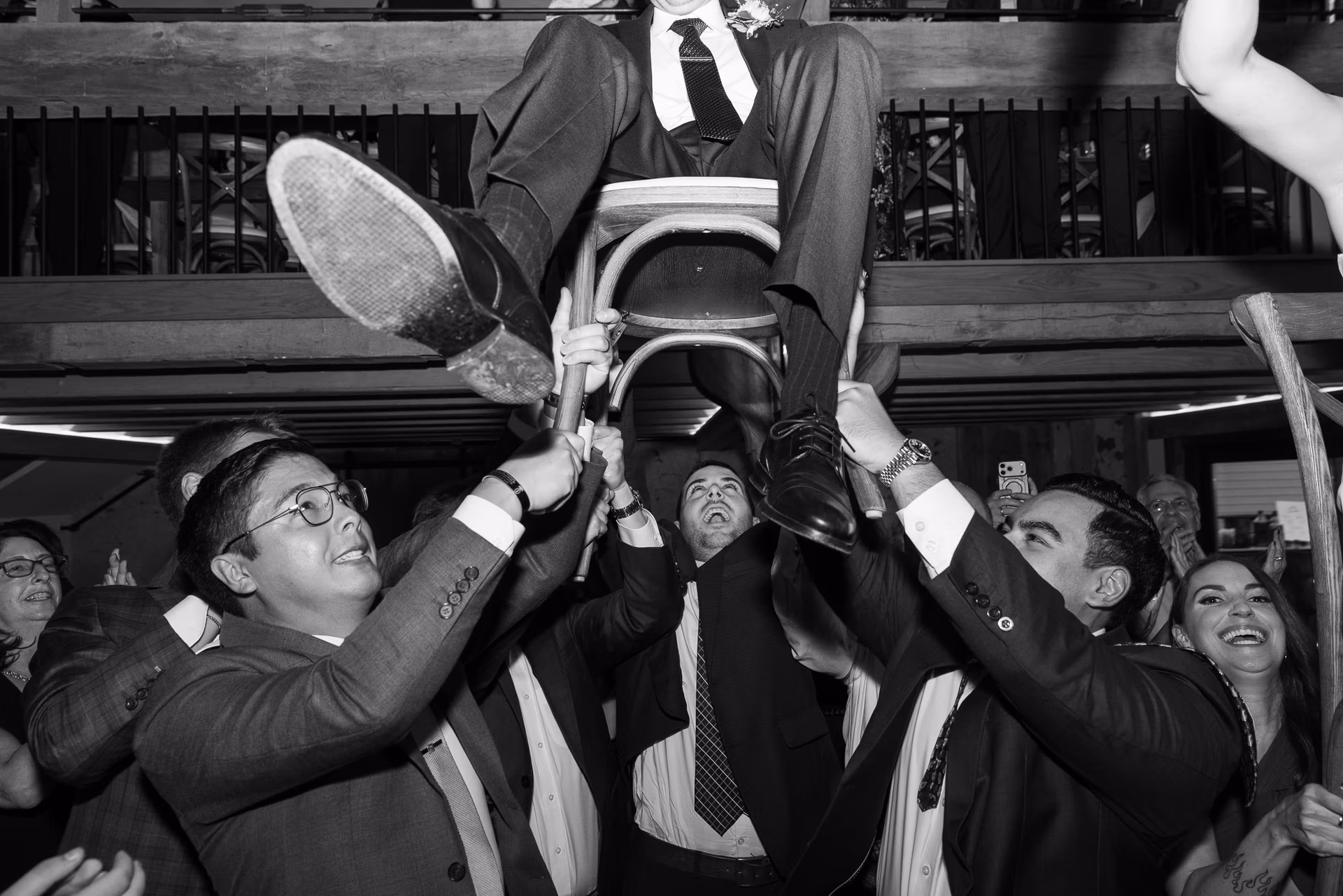The legs and feet of groom in a dark suit dangle as he is lifted on a chair during the Hora with friends and family