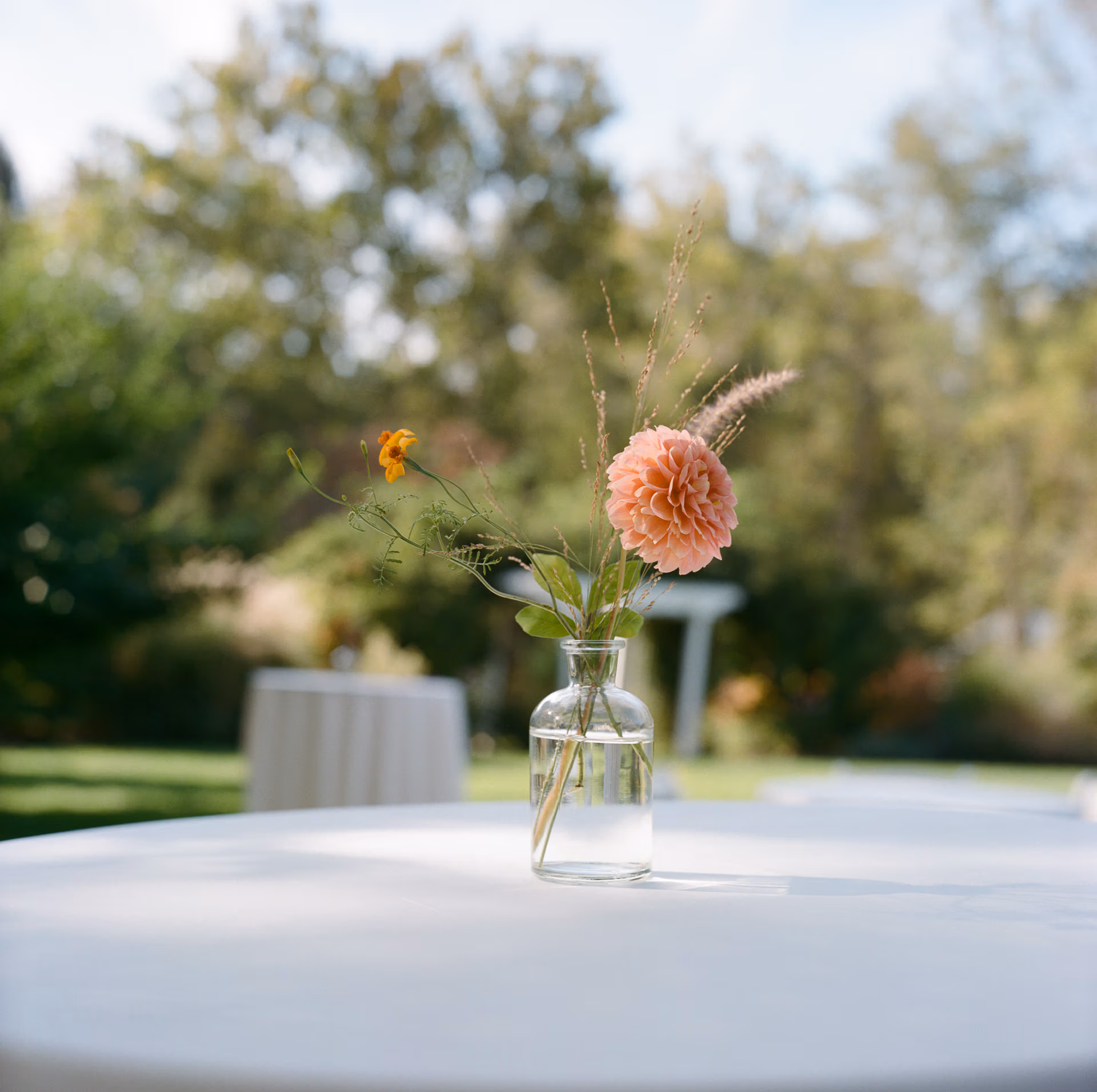 Single coral dahlia stem in a clear bud vase decorates a high top table with a white table cloth during wedding cocktail hour at the Woolverton Inn