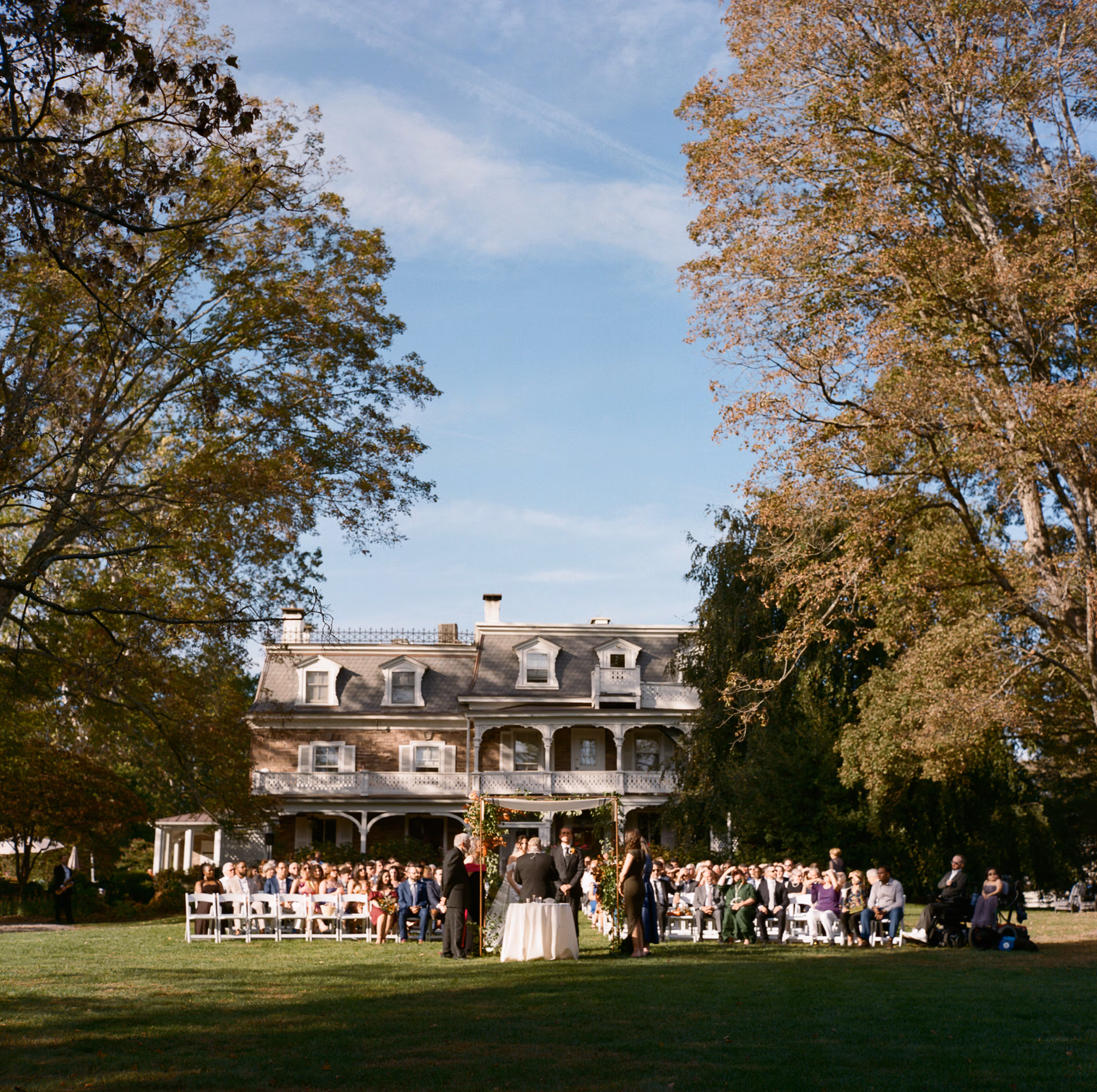 A large gathering of guests watches a wedding ceremony with the historic Woolverton Inn as the backdrop