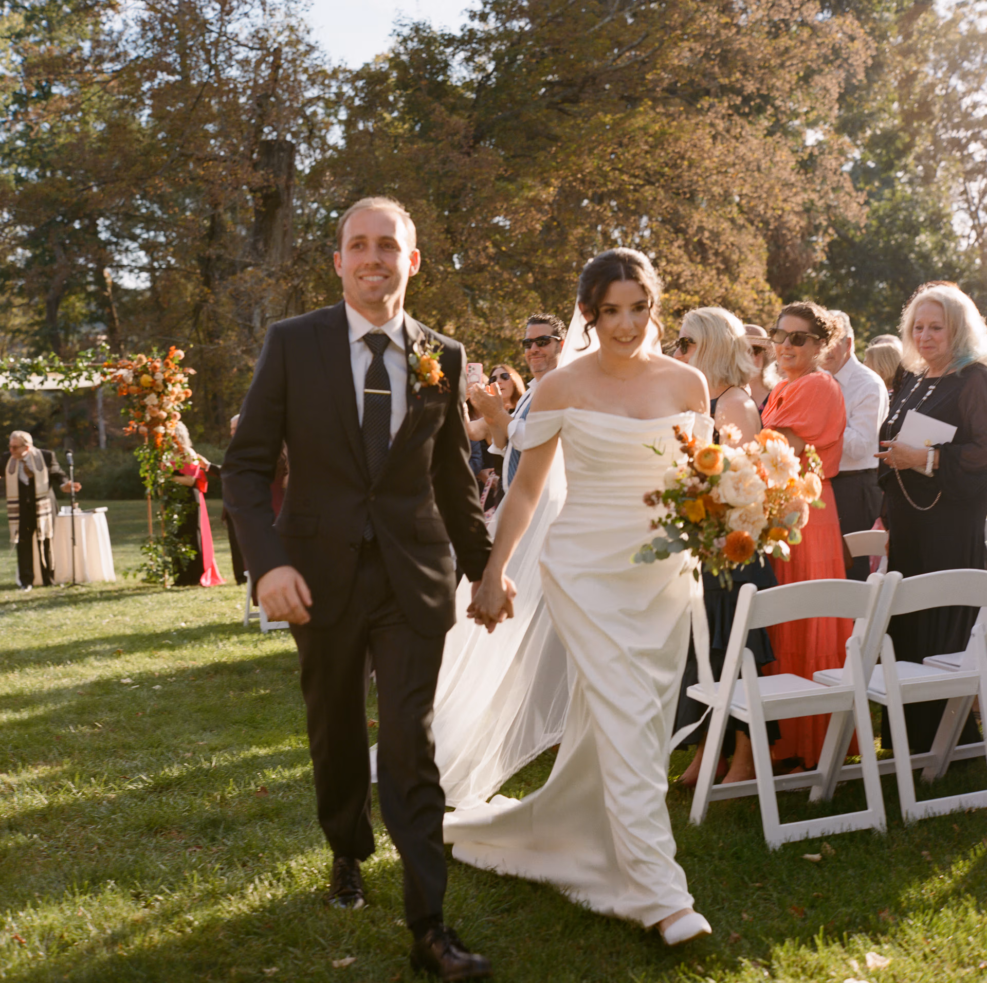 Elegant bride in slim satin off the shoulder gown and long veil and groom in a dark suit and tie walk away after their Woolverton Inn wedding ceremony