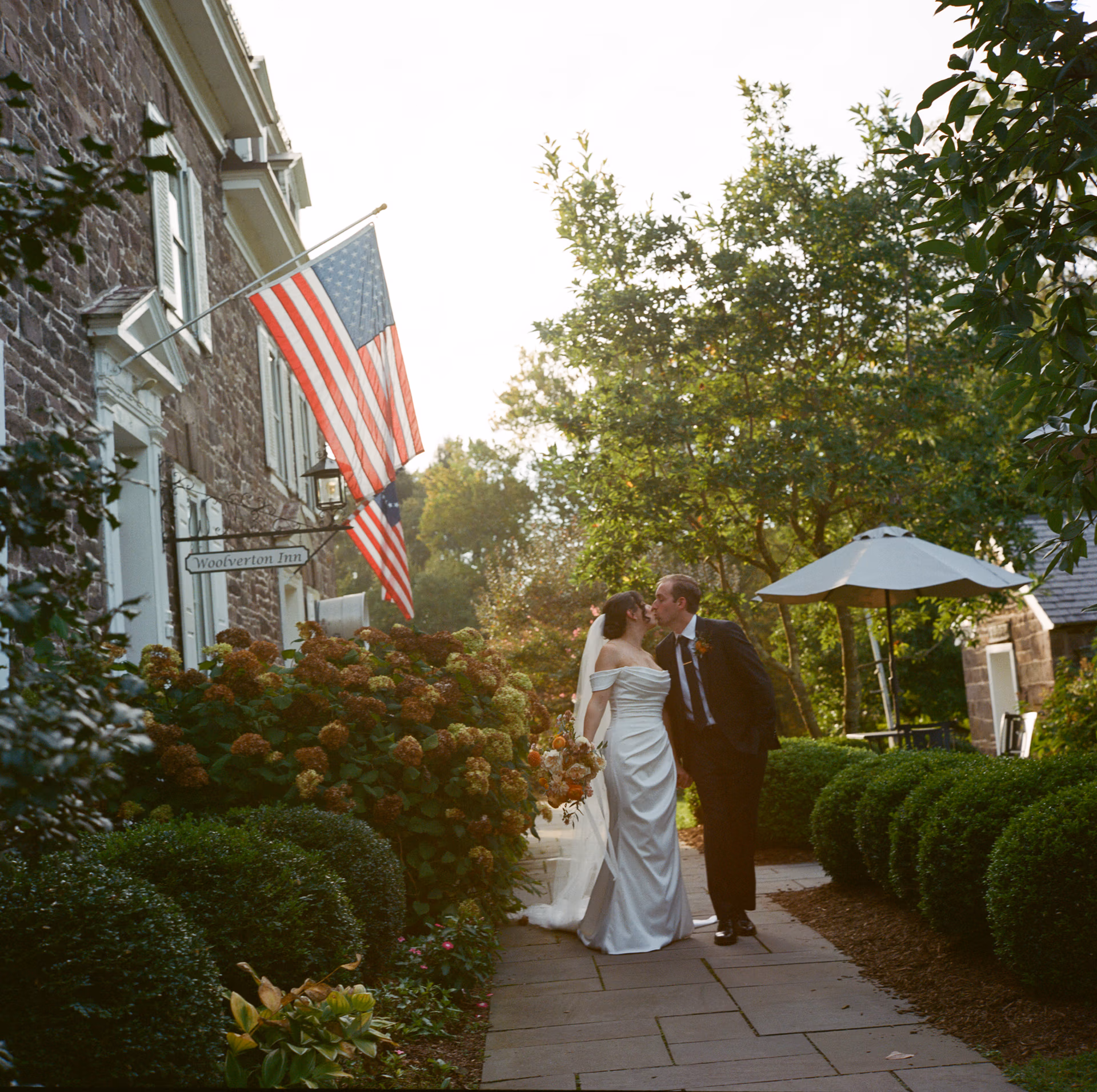 Bride in an elegant gown and long veil and groom in a dark suit kiss on the front walkway with American flag behind them at the Woolverton Inn